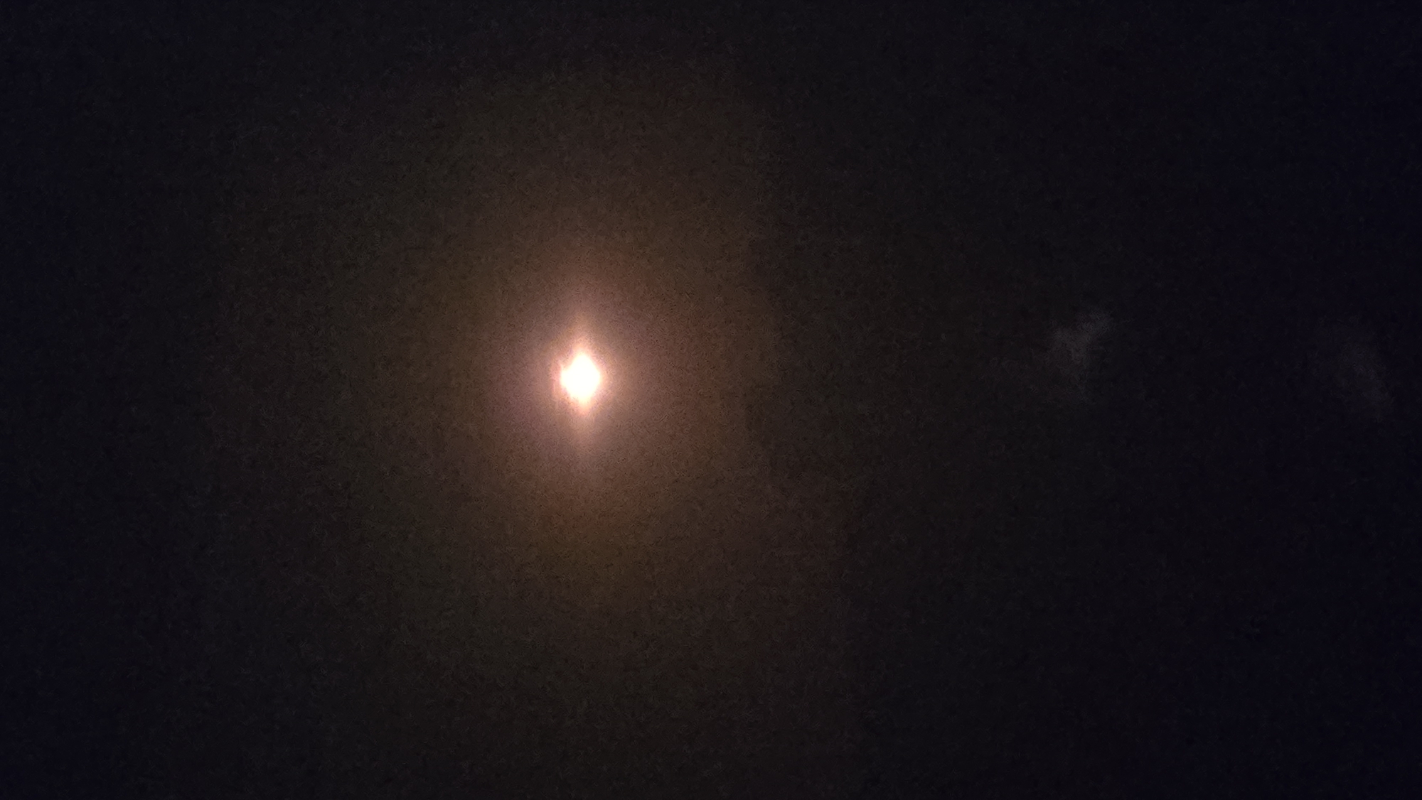 A night sky is visible with a bright, luminous object at the center, likely the moon, surrounded by a soft glow. The background is dark, with subtle hints of lighter, hazy clouds or atmospheric conditions. There are no discernable structures or any indication of the city of Cornellà de Llobregat, Spain. The image is a solitary shot of the night sky and its celestial illumination.