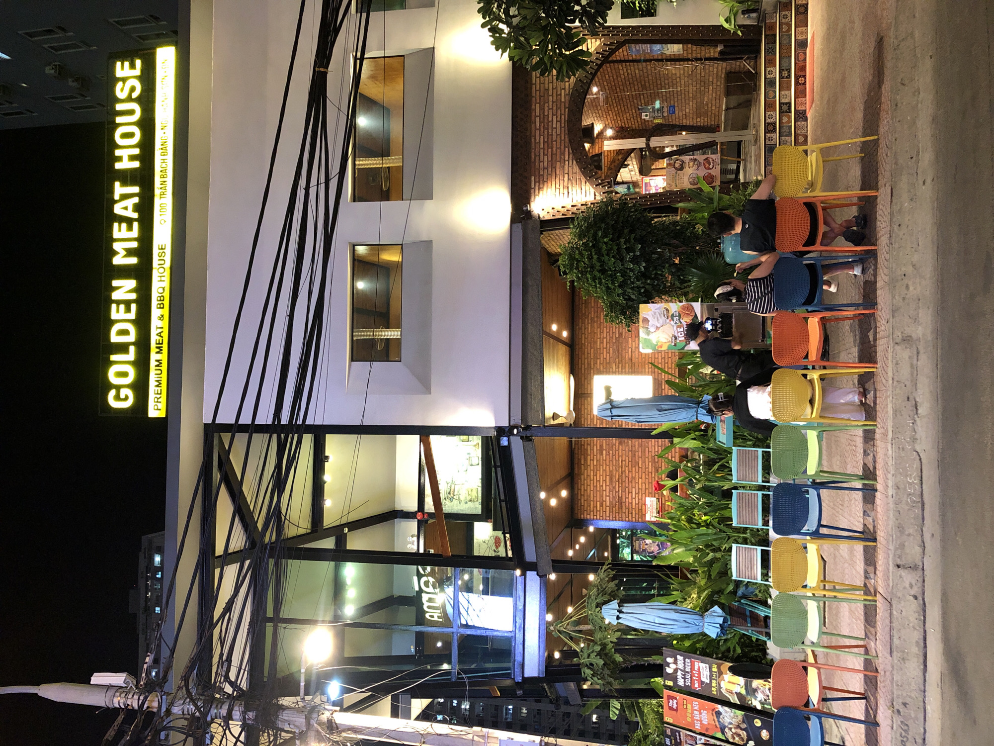 A street-level night view captures the facade of a restaurant named "GOLDEN MEAT HOUSE" in Da Nang, Vietnam. The restaurant, located at 100 Tran Bach Dang, features a modern architectural style with large glass windows and a second-floor balcony. Above the entrance, a bright yellow illuminated sign displays the restaurant's name and "PREMIUM MEAT & BBQ HOUSE." The facade is adorned with brickwork on the lower levels and white painted walls on the upper sections.
In front of the restaurant, a row of colorful plastic chairs, including blue, orange, green, and yellow, are placed along the sidewalk. Several patrons are seated on these chairs. On the left, a server is attending to two customers seated at a small table, both facing away from the camera. Further to the right, another individual is seated facing away from the camera, their back to the restaurant. More empty chairs are visible.
The sidewalk is made of concrete, and beyond the chairs, a paved area with decorative tiles leads up to the restaurant entrance. Several potted plants and umbrellas are strategically placed around the seating area. The street itself is dimly lit, with the restaurant's lights and signage being the primary light sources. Overhead, a network of electrical wires and a utility pole are visible against the dark night sky. A partial view of a multi-story building can be seen in the background.