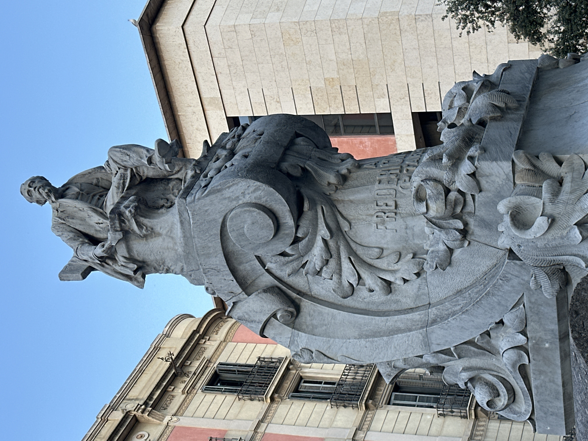 A low-angle, medium shot captures a stone monument in Barcelona, Spain, under a clear blue sky. The monument depicts a seated man with a beard, dressed in a suit and vest. He is looking to his right, with his right hand resting on his lap and his left hand holding a rolled scroll. The monument's base is ornate, featuring carved floral elements and a scroll with the inscription "FREDERIC" visible.
To the left of the monument is a building with light-colored stone facade, pink accents, and balconies with black wrought-iron railings. To the right of the monument is a modern building with a beige stone facade, distinguished by horizontal dark lines. A small bird is perched on the corner of the modern building. A dark green tree is visible in the lower right corner of the frame. The lighting suggests it is either morning or late afternoon.
