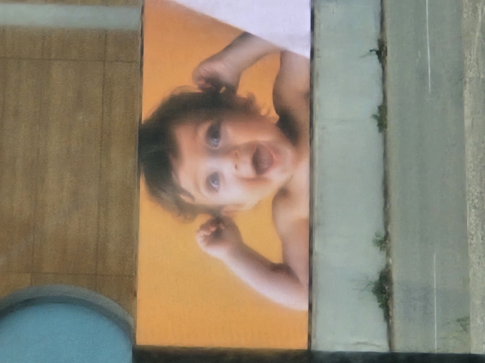 The image displays a rectangular outdoor advertisement featuring a baby, located within the city of Salon-de-Provence, France. The advertisement occupies the central horizontal portion of the frame. The baby, with dark curly hair, is depicted bare-shouldered against a solid orange background, its mouth open wide in an expression of excitement, and both hands raised to the sides of its head near the ears. Its eyes are directed upwards.
Above the advertisement, a light brown, horizontally textured surface is visible, possibly a building facade or awning. A partial circular blue object, possibly a light fixture or decorative element, is visible on the upper left side of this brown surface. Below the advertisement, a light grey concrete wall runs horizontally, with sparse clusters of green vegetation growing along its base. The lowest section of the image reveals a grey asphalt road with faint white markings.
