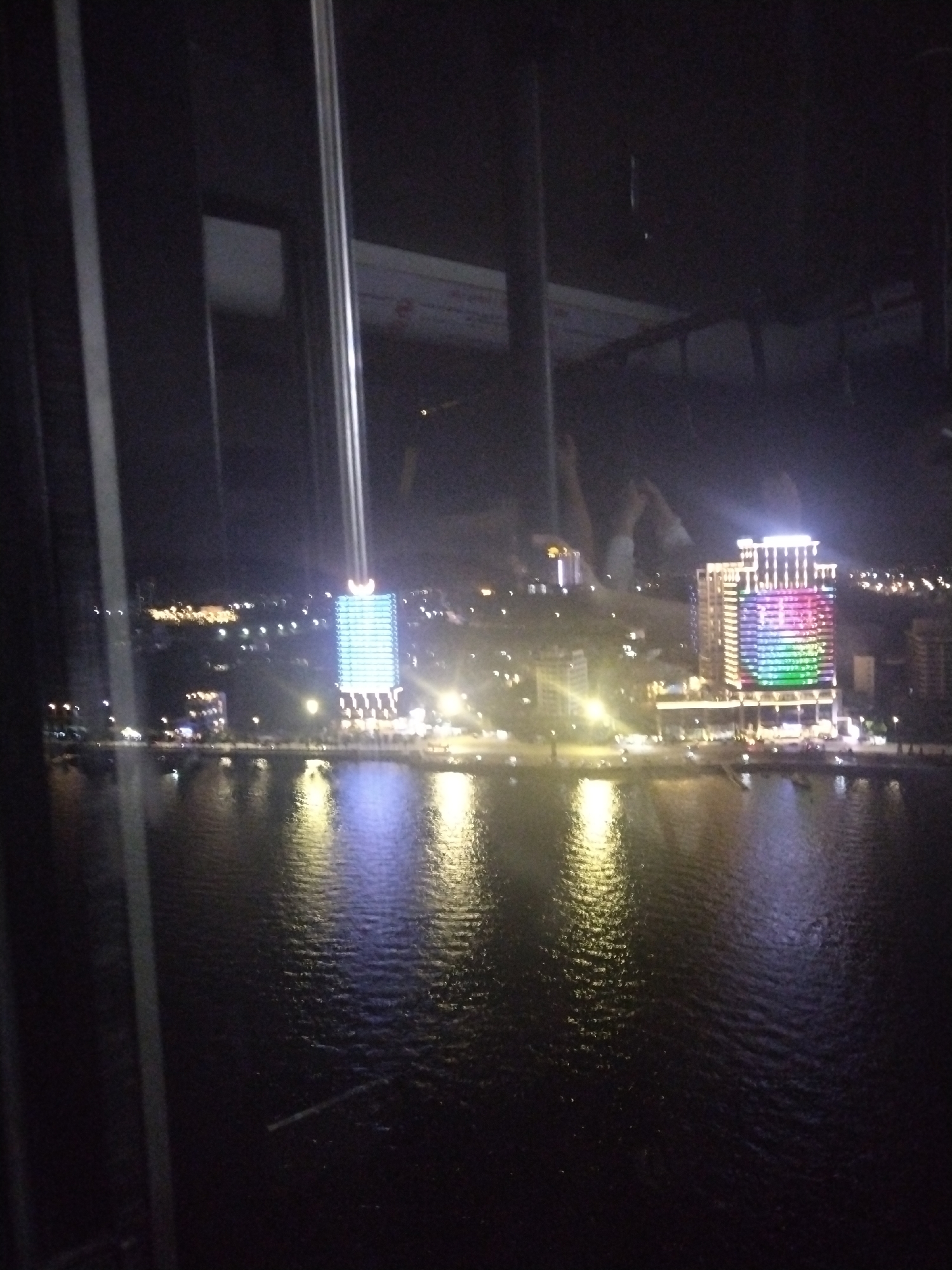 Here's a detailed description of the image:
The image captures a nighttime cityscape reflected in a body of water, likely a river or bay, viewed through what appears to be a window or enclosure.
**Environment:** The overall scene is dark, with the primary source of light coming from the city's illuminated buildings and streetlights. The water surface shows ripples and reflections of the city lights. The city appears densely populated, with many buildings of varying sizes.
**Objects:** Two prominent buildings are visible across the water. One is illuminated with white lights, and the other has a multicolored light display. Numerous other buildings and streetlights line the waterfront, creating a luminous horizon. The image is framed by elements that suggest the view is from within a structure, with window frames visible on the left.
**People:** There's a partial view of at least one person in the reflection. It seems like the individual is holding up their phone to take a picture.
**Location:** The location is likely in a city, given the dense urban landscape, but the specific city is unknown. The image is taken at night.