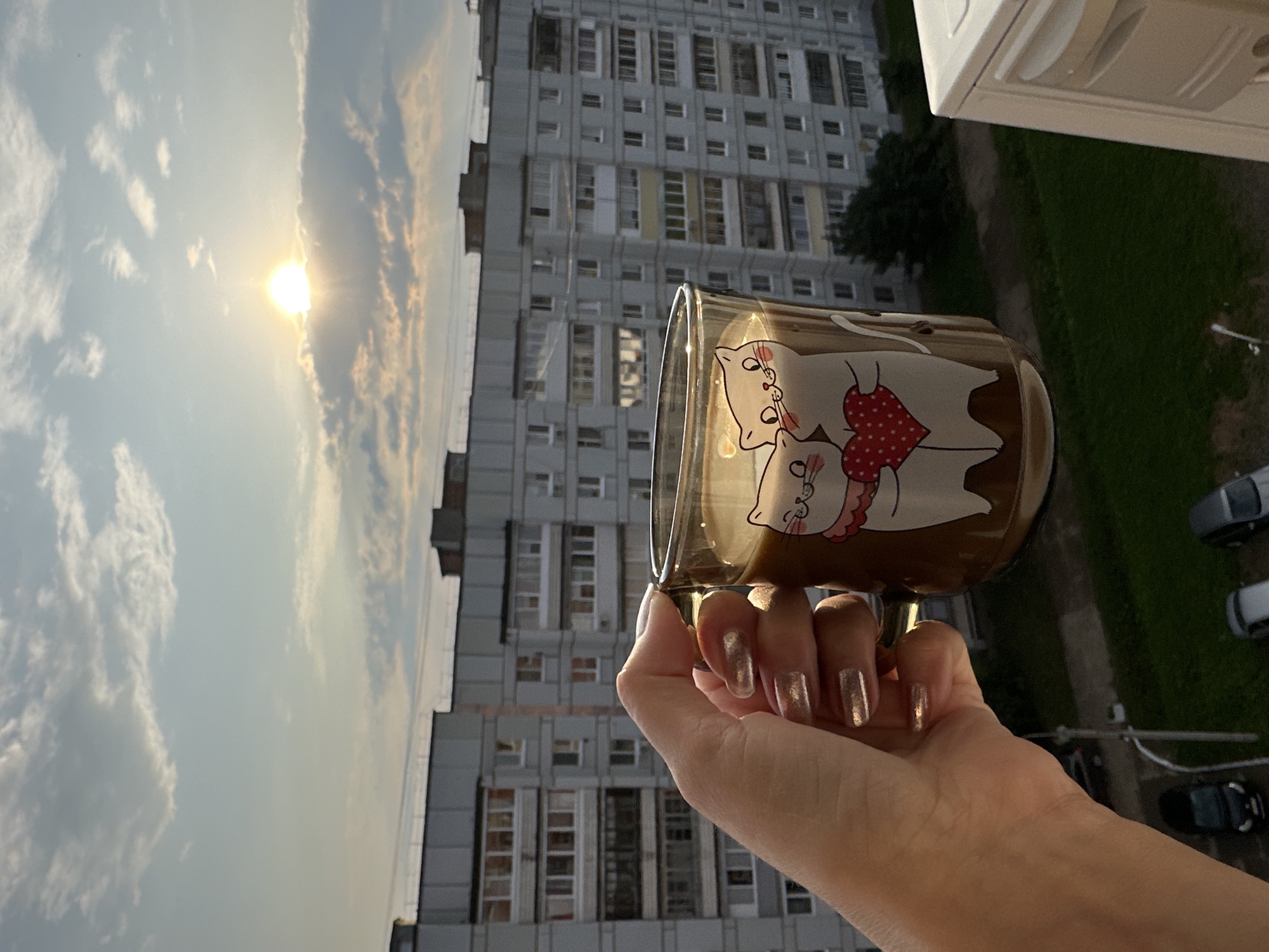 A person's hand with glittery nail polish holds a clear glass mug filled with a light brown beverage, likely coffee or tea, against a backdrop of apartment buildings and a cloudy sky with the sun visible. The mug features a graphic of two cats embracing and holding a red heart with white polka dots. The scene appears to be taken from a balcony or window, overlooking a street with parked cars and a patch of green grass. The apartment buildings are typical of Soviet-era architecture, common in cities like Chaikovsky, Russia. The lighting suggests it is either early morning or late afternoon.
