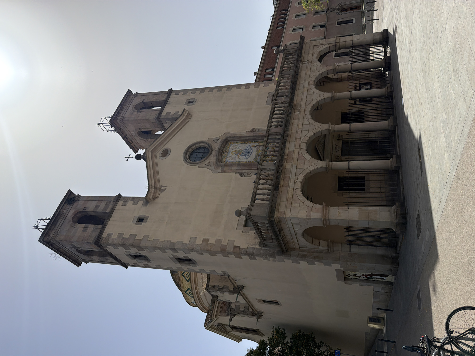 The image shows a wide-angle, eye-level view of the church of Sant Vicenç de Sarrià in Barcelona, Spain, on a clear, sunny day. The church, constructed of light-colored stone and stucco, features a prominent facade with two bell towers flanking a central section. The left tower is slightly more visible and has a more elaborate spire-like structure on top. Below the towers, a large balcony supported by arched colonnades extends across the front of the building. A round stained-glass window is centered above a mosaic depicting a religious figure. The dome of the church, visible on the left side, is adorned with colorful geometric patterns. The surrounding environment is urban, with a paved plaza in front of the church and buildings visible to the right. A bicycle is parked on the left side of the plaza, and a street sign is also present. The sun is high in the sky, casting distinct shadows. No people are visible in the frame.