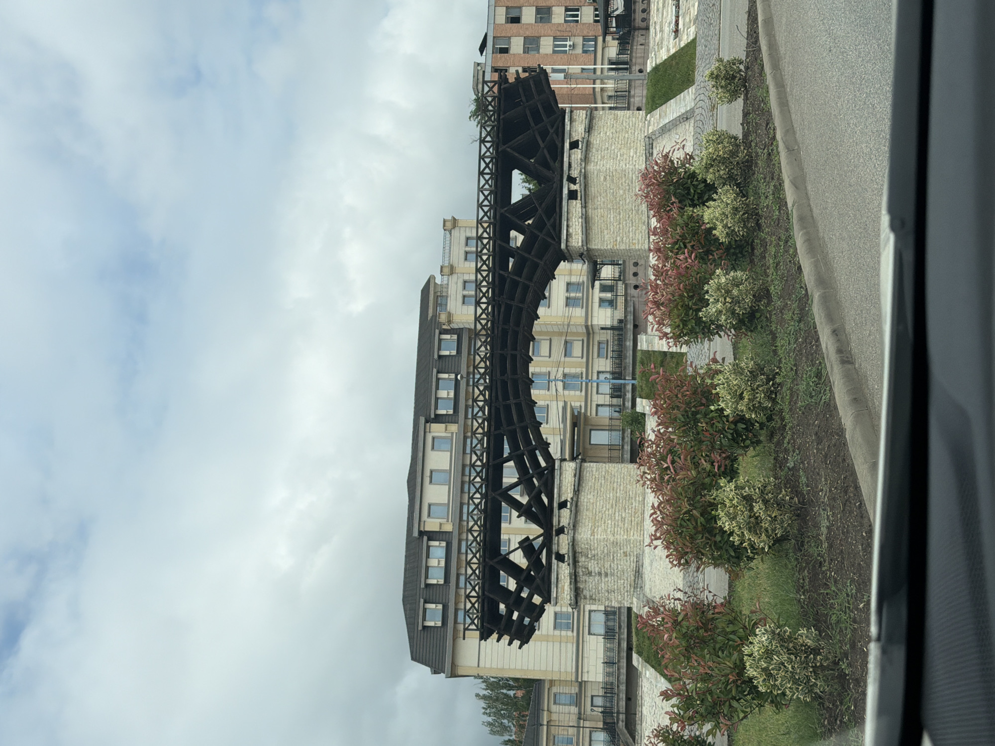 The image, captured from a vehicle, presents a street view in Drobeta-Turnu Severin, Romania, under a cloudy, overcast sky. The central focus is a large, dark, curved architectural structure, resembling a section of a lattice-work bridge, possibly a replica of Trajan's Bridge or the "Eiffel Bridge" located in the city. Its dark, almost charred appearance and intricate grid pattern stand out against the backdrop of urban buildings.
Several multi-story buildings with light-colored facades, numerous windows, and balconies are visible behind and around this structure, suggesting a residential or commercial area. To the right, a paved road or driveway with a concrete curb separates the street from a landscaped strip. This strip features patches of green grass and several decorative bushes with a mix of red and green foliage. The scene is static, devoid of people or discernible activity. The cloudy sky indicates a daytime setting without direct sunlight. No visible text is present in the image.