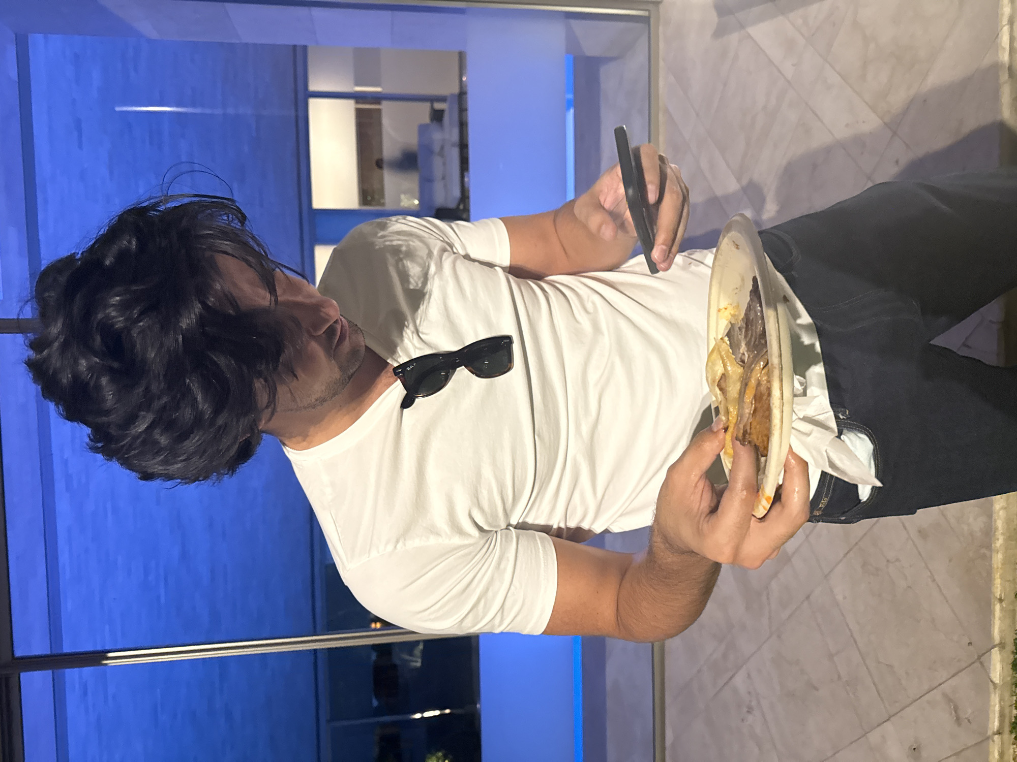 A man with dark hair is standing outside in Mexico City, Mexico, holding a plate of food in his left hand and a cell phone in his right hand. He is wearing a white t-shirt and dark pants. Sunglasses are placed on his t-shirt. The area appears to be a terrace or outdoor area with marble flooring, with a glass wall or large window in the background reflecting an interior space. Outside the window, there is greenery indicating an outdoor environment. The setting suggests a modern or upscale urban environment. The man appears to be taking a photo of his food.