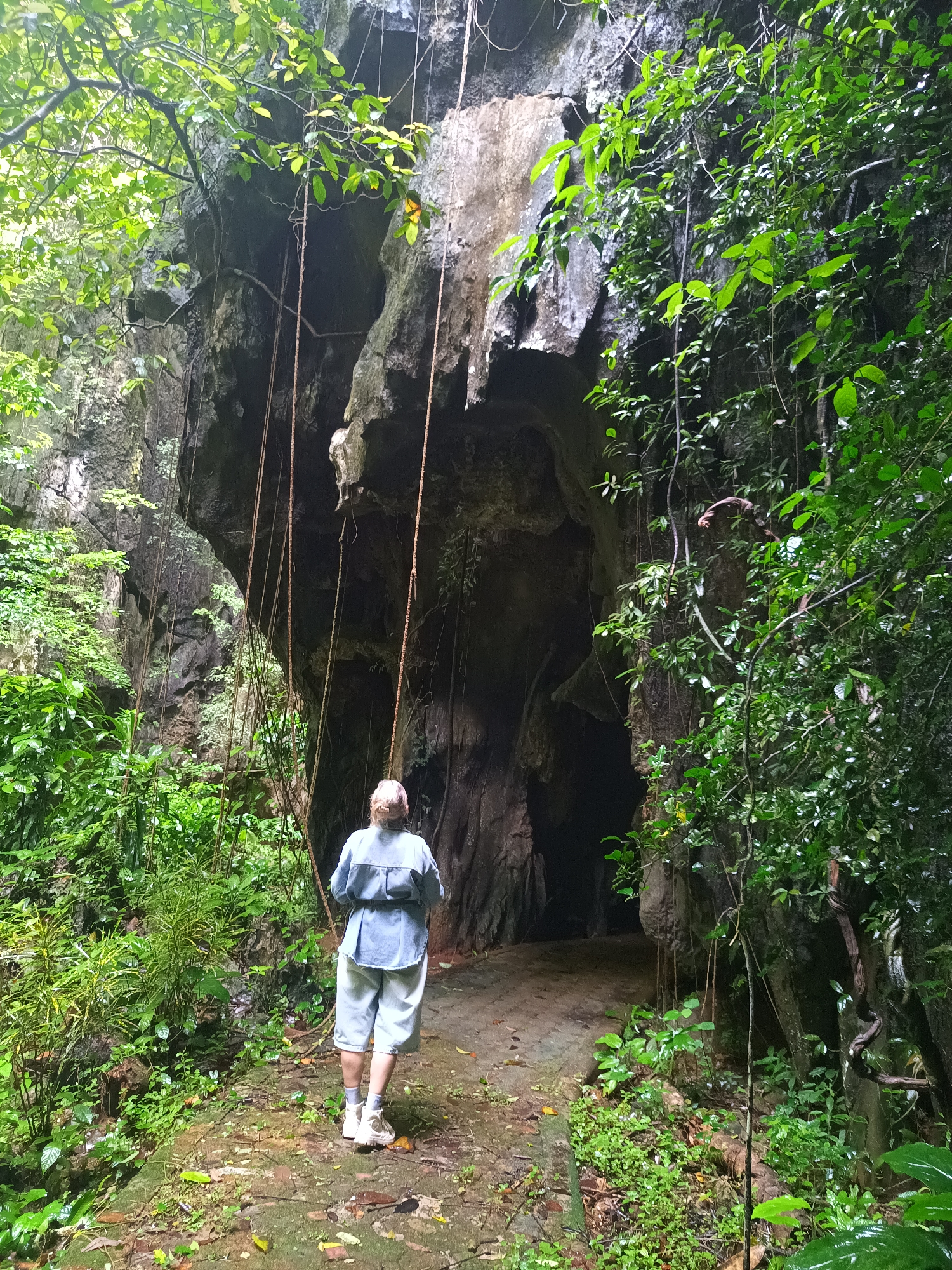 A woman stands at the entrance to a dark cave, located in Ban Pak Ra, Thailand. She has short blond hair, is wearing a light blue shirt, light blue shorts, white socks, and white shoes. The ground is covered with green vegetation. The cave opening is surrounded by large rocks and vines. The overall environment is lush and green.