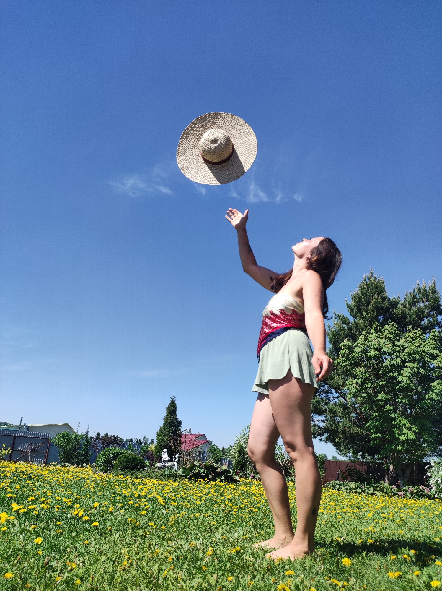 A woman stands barefoot in a field of dandelions beneath a clear blue sky, seemingly reaching for a straw hat suspended in the air above her. She is wearing a patterned top and light green shorts. Behind her are trees and a view of buildings in the distance. The image was taken in a rural area with houses in the background. Based on the location, I can infer that this image was taken in the city of Фоки, Russia.