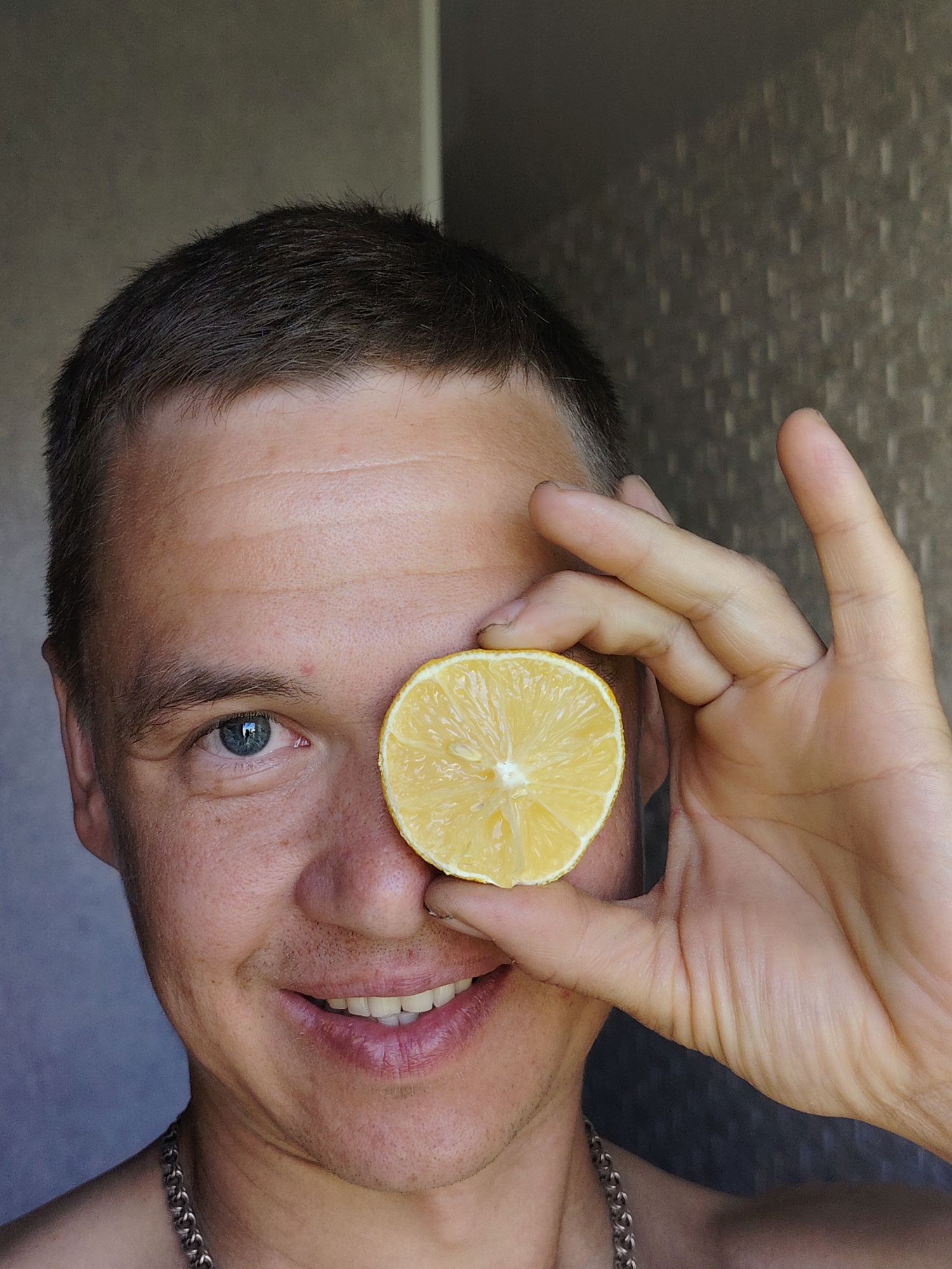 A close-up, brightly lit selfie depicts a man holding a halved lemon over one eye. The man has short, dark hair and light skin with freckles. He is smiling, revealing his teeth. He is wearing a silver chain necklace. A portion of his bare shoulder is visible. The background is a textured gray wall. The location is not discernible from the image. Therefore, it's not possible to determine if the image was taken in Chaikovsky, Russia.