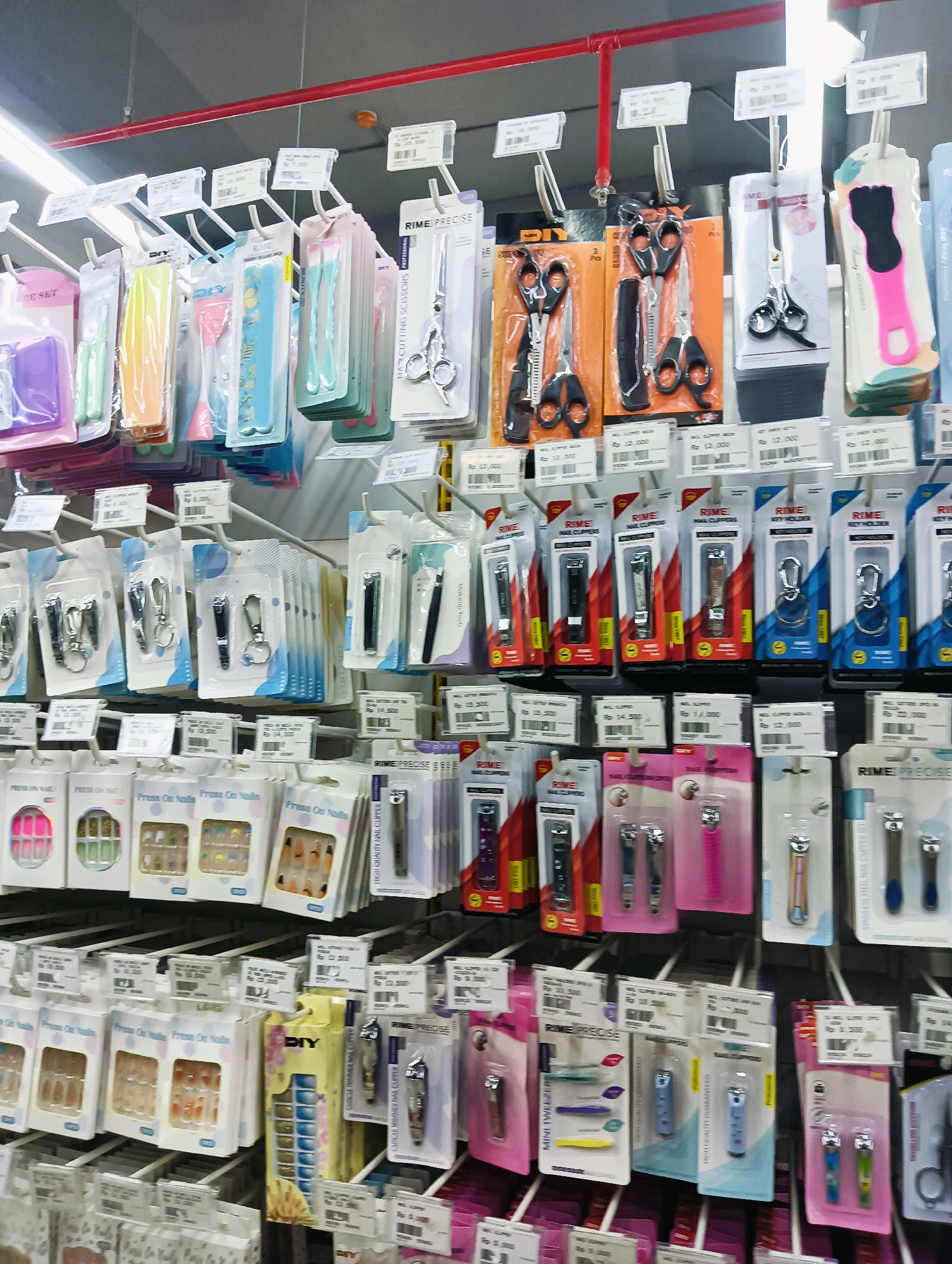 The image depicts a retail display of personal grooming and beauty products, likely in a store in Batam, Indonesia. The items are neatly arranged on shelves and hanging displays, primarily featuring nail clippers, scissors, tweezers, and press-on nails. The lighting appears to be artificial, consistent with an indoor retail environment. The products are mostly packaged in clear plastic and cardboard, with brand names and prices visible on some labels. The overall impression is one of a well-stocked and organized merchandise display.