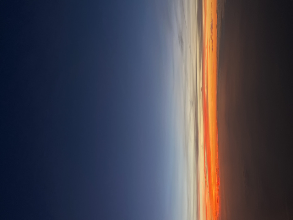 The image displays an aerial view of the sky at either sunrise or sunset. The upper portion of the frame is a deep, dark blue, transitioning downwards into a lighter blue, followed by subtle bands of grey and white clouds. Below these, a prominent, vibrant horizon line features intense orange and red hues, indicative of atmospheric scattering of light. The lowest portion of the image consists of a dense, dark cloud layer, appearing as an indistinct, undulating mass that extends across the bottom third of the frame. No ground features, man-made structures, people, or identifiable objects are present. The scene captures atmospheric conditions only. As an aerial view, no specific location within the city of Freising, Germany, can be identified from the visible details.
