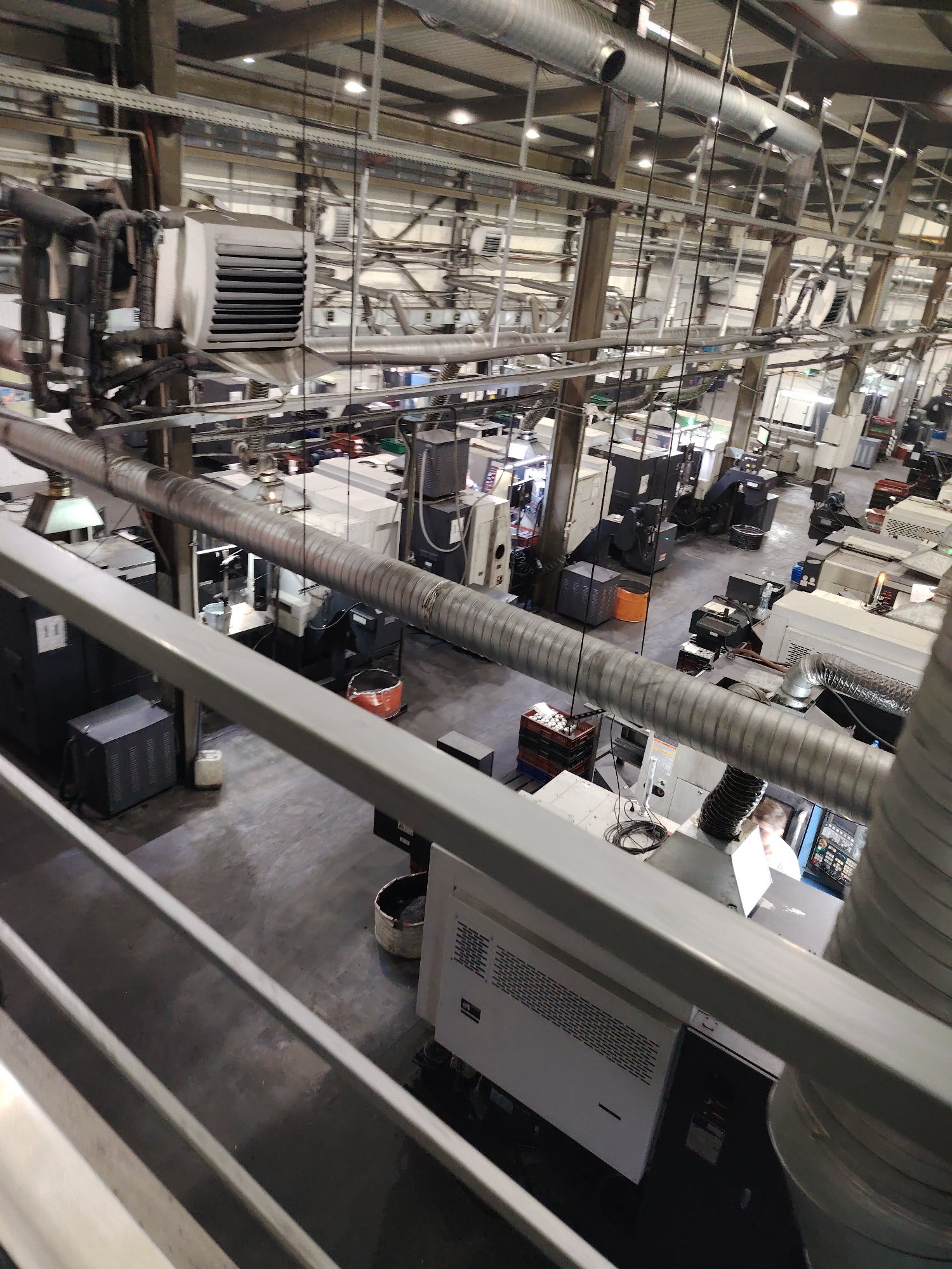 The image depicts the interior of a large industrial facility, possibly a manufacturing plant, in Noviy, Russia. The location is characterized by numerous pieces of heavy machinery, likely Computer Numerical Control (CNC) machines or similar automated manufacturing equipment. The machinery is arranged across a wide, open floor, with a grey, industrial-grade floor surface. Overhead, there is an extensive network of pipes, ventilation systems, and electrical conduits suspended from the ceiling, typical of industrial environments. The lighting appears to be provided by rows of fluorescent or LED fixtures, creating a bright, utilitarian atmosphere. In the foreground, a railing or platform suggests the photo was taken from an elevated position. A person, seemingly an operator, is seen interacting with one of the machines. The environment is utilitarian, focusing on functionality and operational efficiency rather than aesthetic appeal.