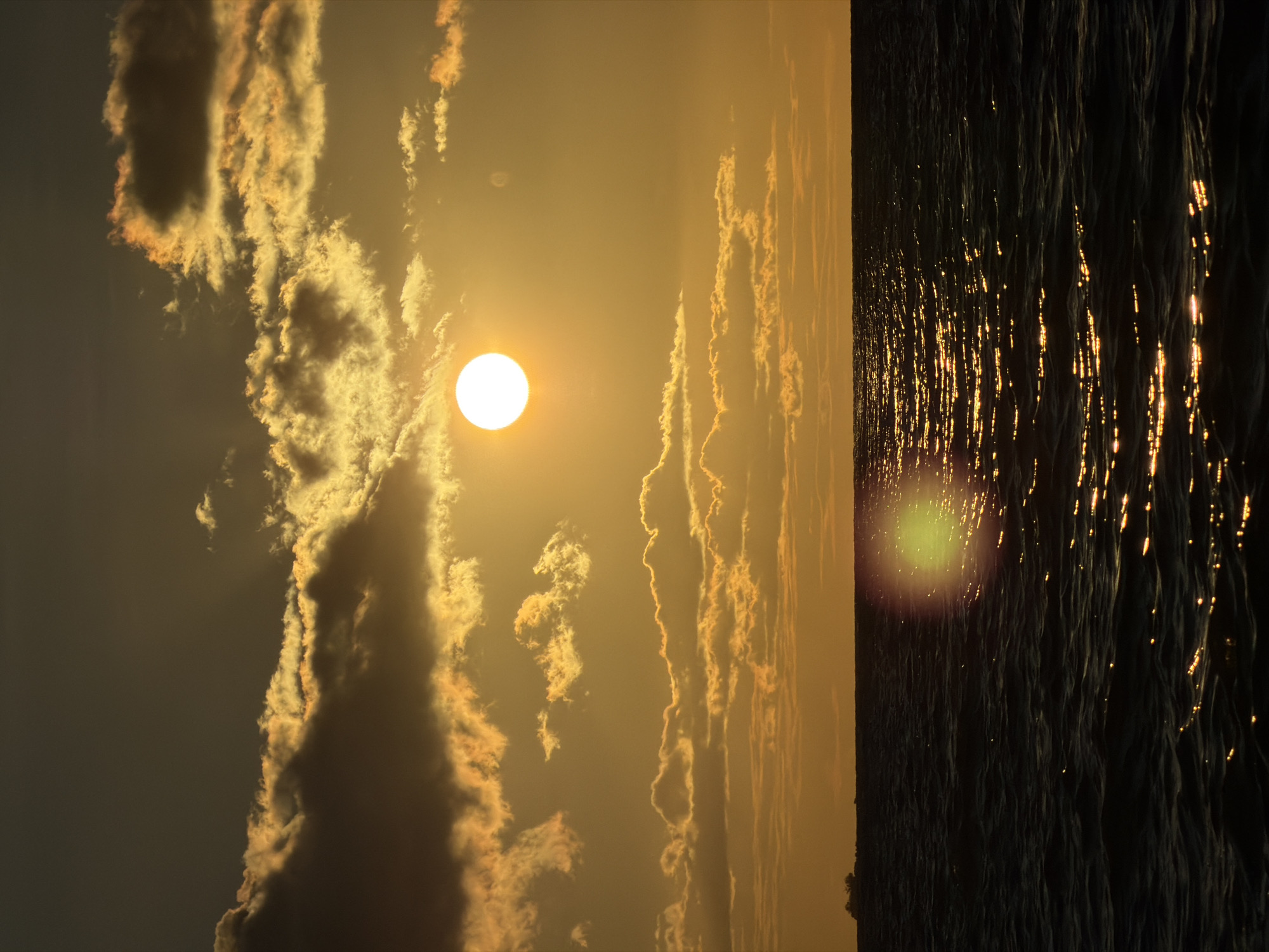 The image depicts a sunset over the ocean. The sky is a gradient of orange and yellow, with scattered clouds that are illuminated by the setting sun. The sun itself is a bright, white orb positioned in the upper right quadrant of the sky. The ocean is dark and appears to be choppy, with reflections of the sun shimmering on the water's surface. In the far distance, on the horizon, a small landmass or ship is faintly visible. There are no people or specific identifiable objects within the scene besides the natural elements of the sky and sea. The location is stated to be Baan Bo Phut, Thailand, implying a coastal setting.
