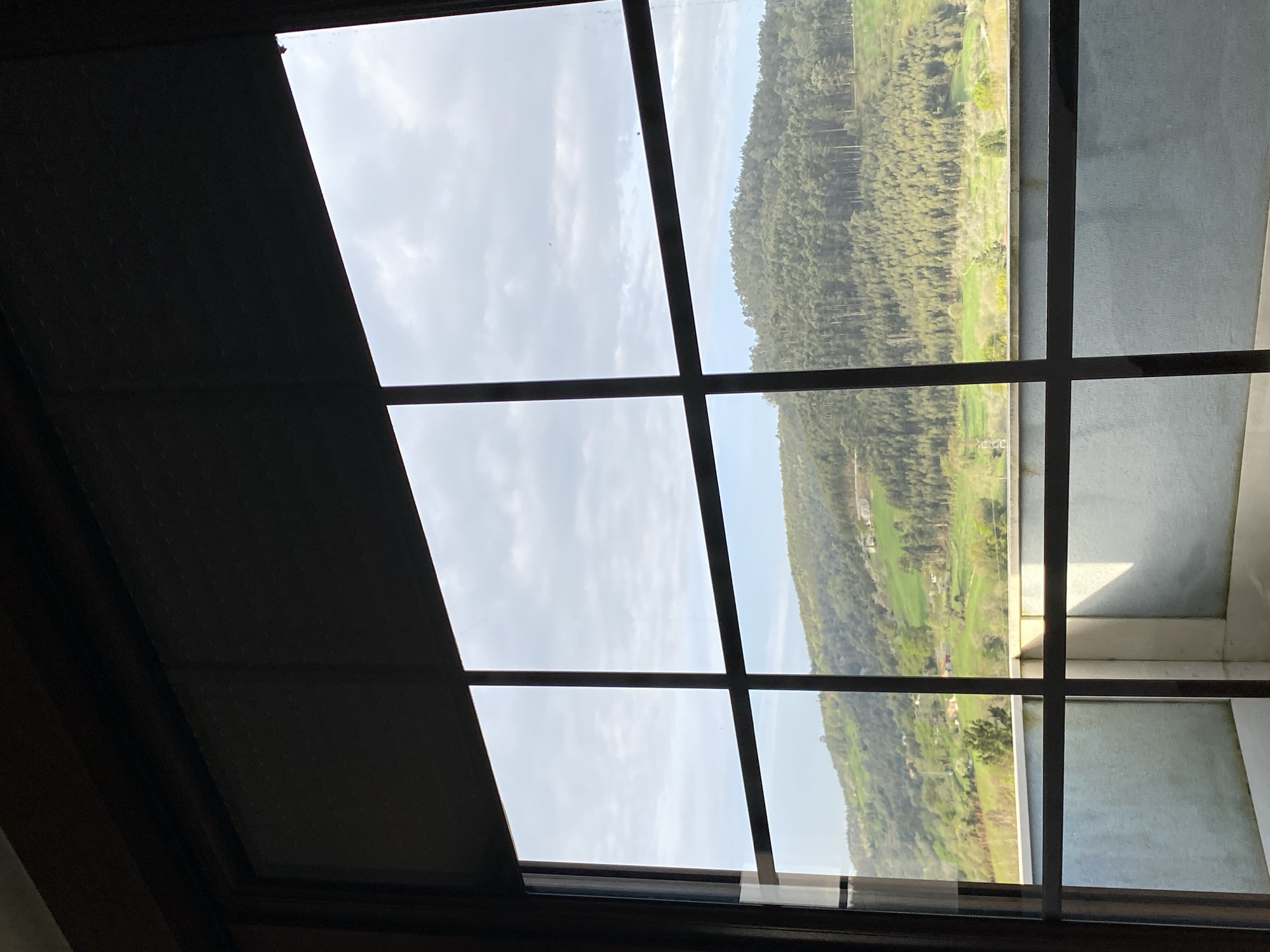 The image is taken from inside a building looking out through a window with a grid pattern. The view outside is of a landscape featuring a hillside covered in trees and green fields. The sky is overcast with clouds. A portion of the window is obscured by a lowered blind or shade. The location is inferred to be Villaviciosa, Spain, based on the prompt. The interior appears to be a room with a simple, undecorated wall visible on the right. There are no people visible.