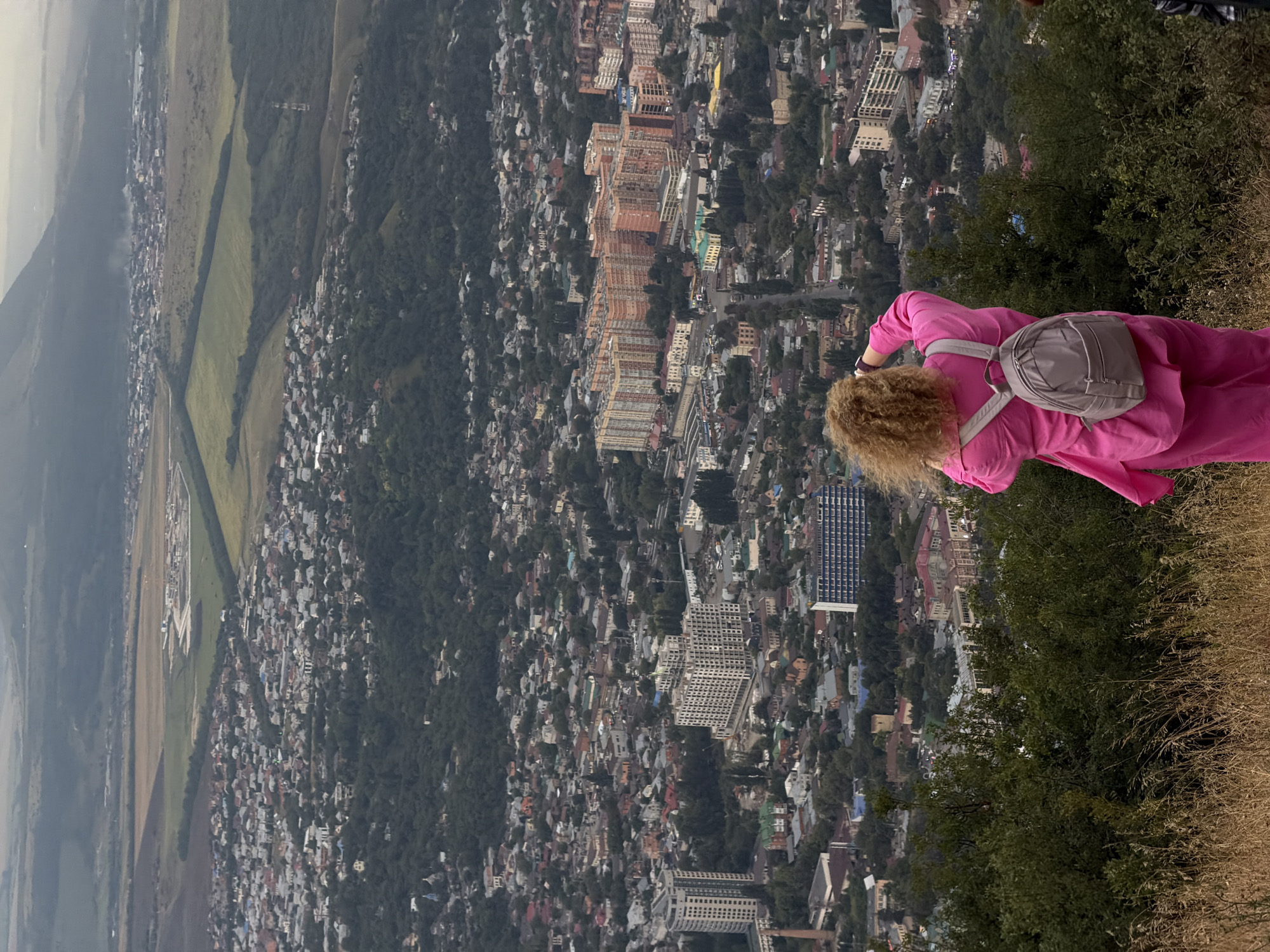 In this image, a woman with curly blonde hair stands on a grassy, elevated vantage point, facing away from the camera and towards a sprawling cityscape. She is wearing a bright pink outfit and a light gray backpack. Her hands are raised to her face, as if taking a photograph or adjusting something. The city below is densely populated with a mix of residential buildings, some appearing to be apartment complexes, and smaller houses. Green trees are interspersed throughout the urban landscape, and in the distance, rolling hills and mountains are visible under an overcast sky. The terrain in the foreground is dry and covered in tall grass, suggesting a natural, undeveloped area overlooking the city. The overall impression is one of a scenic overlook of Goryachevodsky, Russia, with the woman capturing the expansive view.