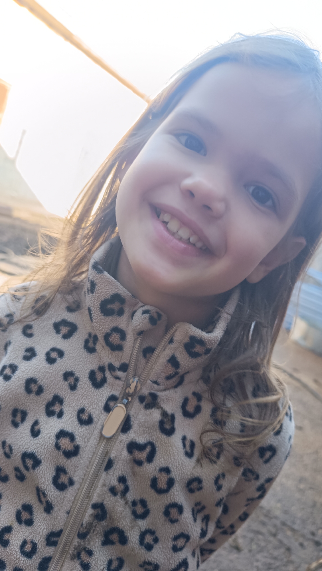 An overhead, close-up shot shows a young girl smiling. She is wearing a tan-colored fleece jacket with black leopard spots and a silver zipper. Her hair is brown and falls around her shoulders. The background is blurry and consists of muted colors, suggesting an outdoor setting. The lighting is bright and appears to be natural, possibly from the sun. The image quality is soft and somewhat grainy. The girl's face fills most of the frame, and her eyes are dark brown. Her teeth are visible as she smiles. The focus of the image is on the girl's face and upper body. The overall impression is one of warmth and happiness.