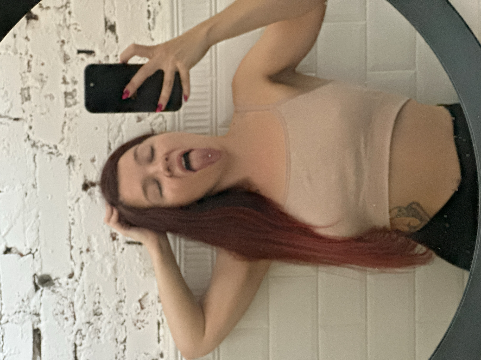 The image is a mirror selfie taken by a woman with long, reddish-brown hair. She is wearing a light beige crop top and black bottoms. Her eyes are closed, and her tongue is sticking out, showing a tongue piercing. She is holding her right hand to her head and her left hand is holding a black smartphone to take the picture. The background appears to be a white tiled wall and a white brick wall. The woman has a tattoo on her lower abdomen. The image was taken in Vladimir, Russia.