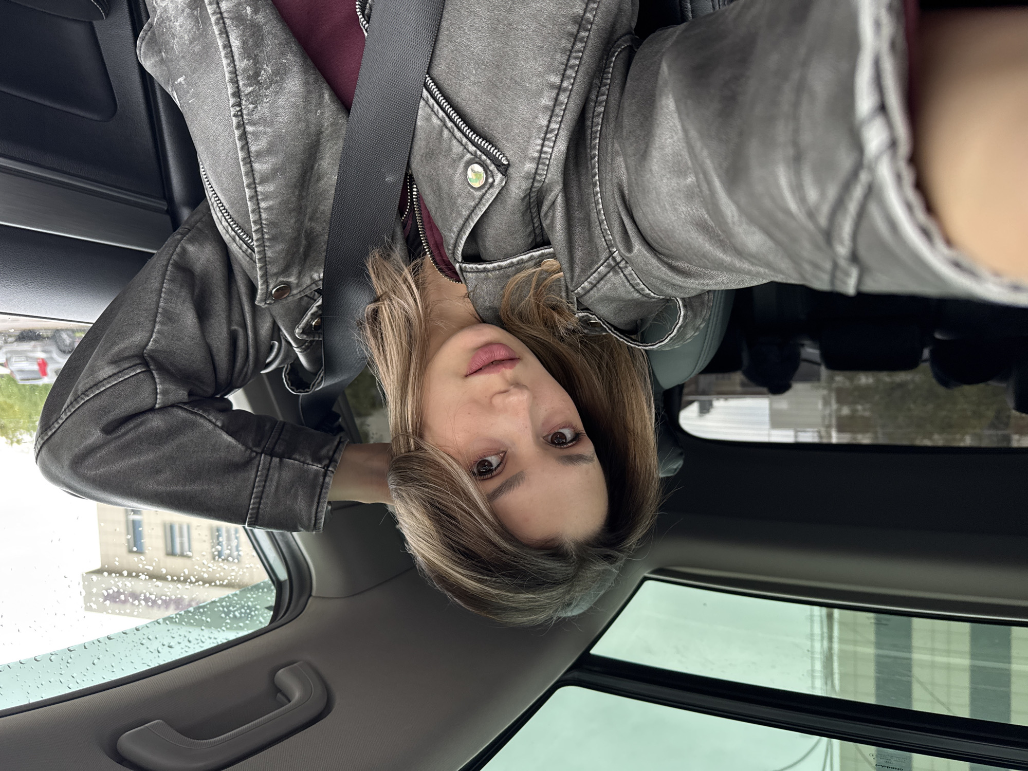 A woman is photographed from a low angle, seemingly inside a car. She has shoulder-length, light brown hair with blonde highlights and is wearing a grey, slightly distressed leather jacket over a maroon top. A black seatbelt crosses her torso. The interior of the car is visible, including the grey ceiling, the window, and the black trim. Outside the car window, there is a building with multiple windows and rain droplets on the glass. Another building is visible through the sunroof. The location is Unknown, Unknown.