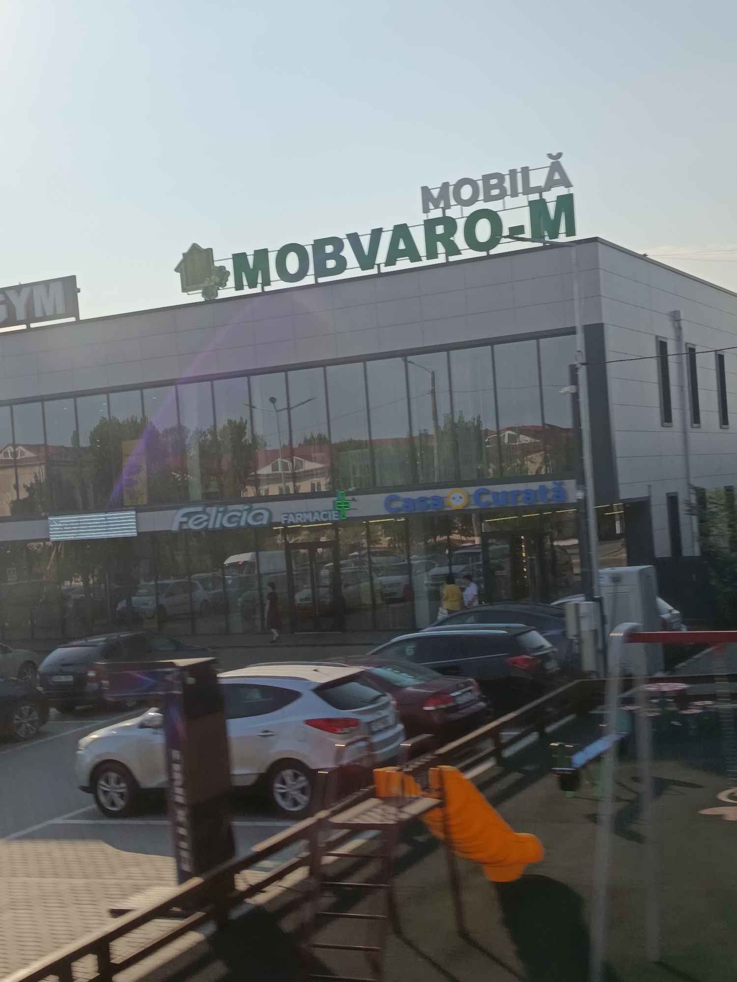 The image shows the facade of a commercial building with large glass windows. Above the windows, the words "MOBILĂ MOBVARO-M" are visible in large, green, capital letters, with "MOBILĂ" in smaller white letters above. To the left, the word "GYM" is visible in white capital letters on a dark background. Through the glass windows, reflections of trees and buildings are visible, as well as several parked cars. On the ground floor of the building, signs for "Felicia FARMACIE" and "Casa Curată" are visible. There are several cars parked in front of the building, and a playground with an orange slide is visible in the foreground to the right. The scene appears to be in an urban or suburban setting, possibly a shopping center or business park, located in Cahul, Moldova. The sky is clear and bright, suggesting it is daytime.