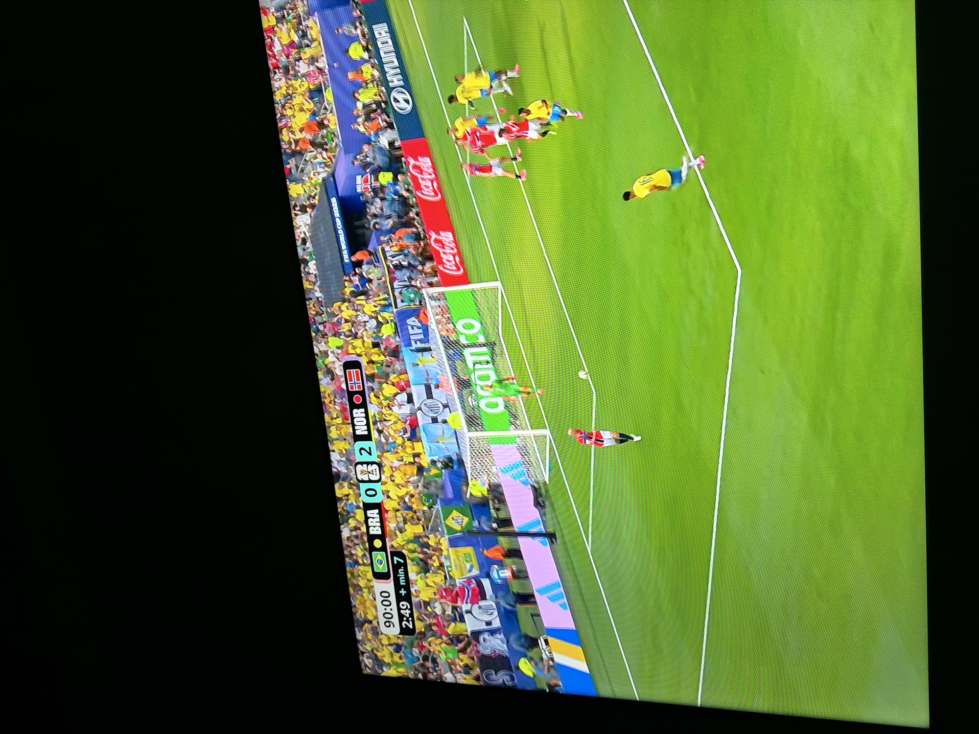 This is a low-angle, eye-level shot of a football match displayed on a television screen. The screen displays a game with the score "BRA 0 - 2 NOR". The time shown is "90:00 + min. 7". The pitch is green with white lines. Several players are visible on the field. Two players in yellow jerseys are on the right side of the screen, and a player in a red jersey is in the center foreground. In the background, a crowd of spectators wearing yellow shirts can be seen. Advertisements for "FIFA WORLD CUP 2026", "Hyundai", "Coca-Cola", and "aramco" are visible along the sidelines. The image is tilted, and the bottom edge of the television screen is visible at the very bottom of the frame, with a black border. The photograph appears to have been taken in the city of Ognianovo, Bulgaria, as indicated by the context of the query.