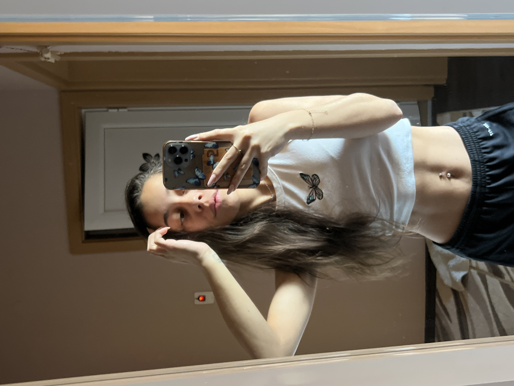 A young woman is captured in a mirror selfie, seen from the chest up, with the image rotated 90 degrees counter-clockwise. She has long, dark brown hair and a relaxed expression. She holds a smartphone with a butterfly-themed case, likely an iPhone due to the triple camera array, to take the picture. She is wearing a white ribbed crop top with a colorful butterfly design on the left side and black athletic shorts with the text "DRY-FIT" visible on the right leg. A belly button piercing with two silver balls is visible. She wears a thin golden ring on her right hand (reflected left) and a delicate golden chain bracelet on her left wrist (reflected right).
The background, reflected in the mirror, shows an indoor setting. There's a wooden frame visible at the top and sides, likely part of the mirror's border or room decor. A white door frame is visible behind her head, and further back, a wall with a light switch or outlet featuring a red indicator light is seen. In the lower right of the reflection, a patterned fabric, possibly curtains or bedding, is visible. The lighting appears to be artificial, typical of an indoor environment.