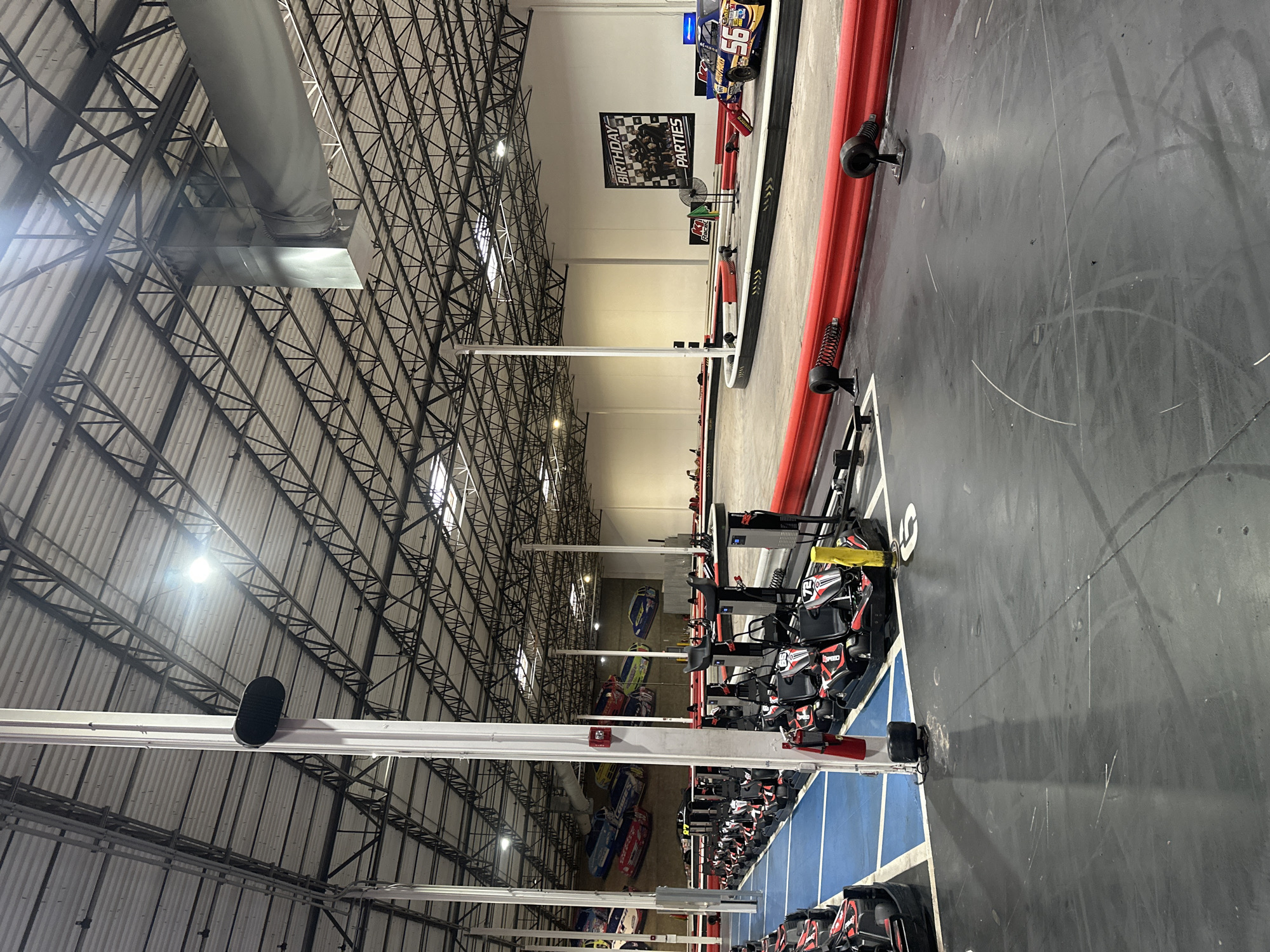The image depicts the interior of an indoor go-karting facility in East Lake-Orient Park, United States of America. A series of go-karts are lined up in a blue-marked starting area, with additional karts visible further down the line. The track itself is gray with red safety barriers. A sign promoting "Birthday Parties" is visible in the distance. The ceiling is a high, exposed metal framework with lighting fixtures.