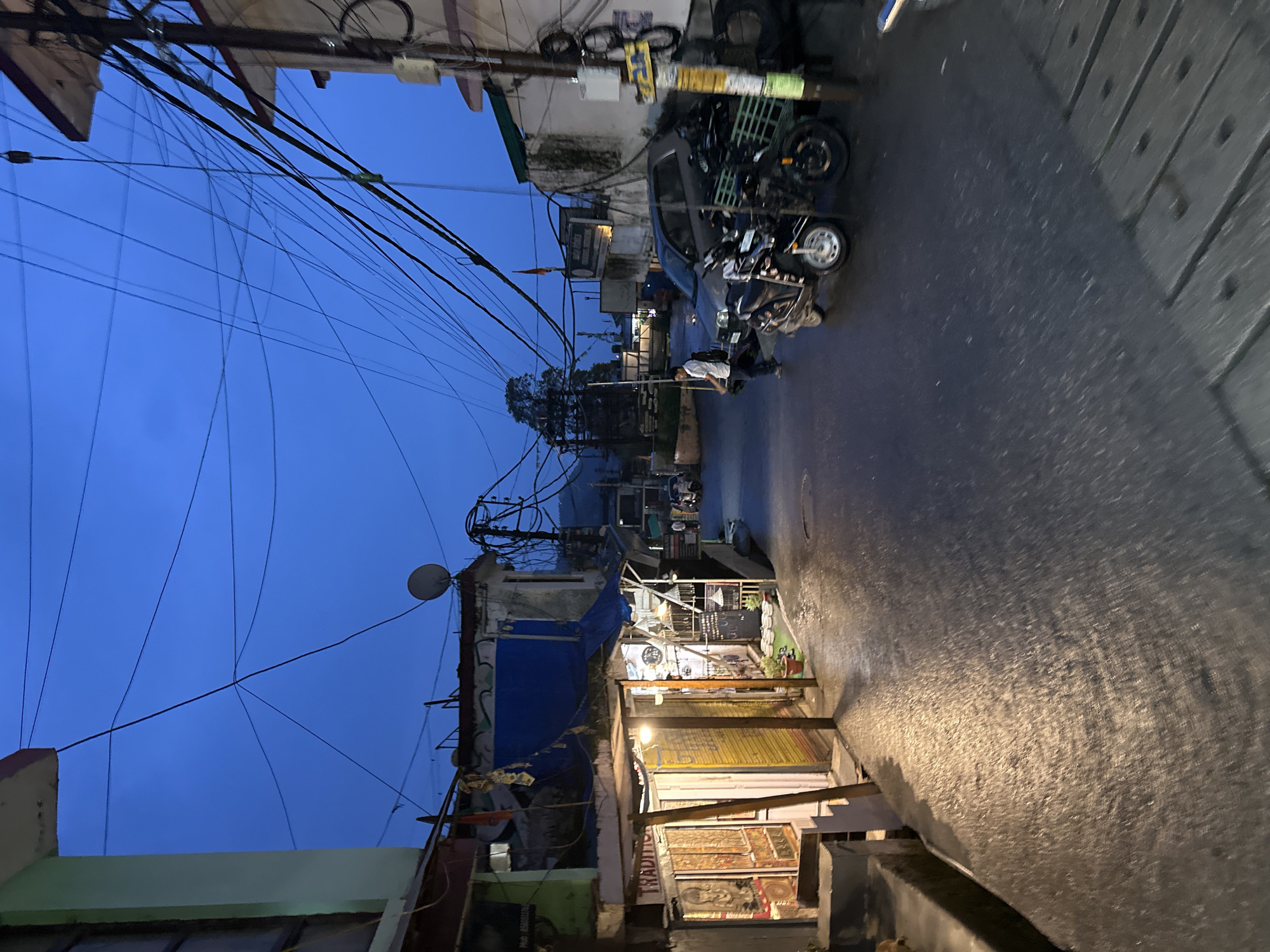 The image depicts a street scene in what appears to be a bustling market area at dusk or early night. The sky is a deep blue, indicating low light conditions. The street is wet, reflecting the ambient light from street lamps and shop fronts, suggesting recent rain or cleaning.
On the left side of the street, there is a row of shops with open fronts. One shop has a sign that reads "TRADITIONAL." The shops display various goods, including what look like decorative items and textiles. Lights illuminate the interiors and awnings of these shops.
The street itself is paved and appears to be a two-way road with parked vehicles and moving traffic. In the middle ground, a person in a white shirt and dark pants is walking on the street, carrying a backpack. Several motorcycles and a car are parked or stopped on the road. One motorcycle is in the foreground, parked at an angle.
The street is lined with buildings on both sides, featuring multiple stories. A complex network of overhead electrical wires and cables crisscrosses the sky above the street, a common feature in urban environments. A satellite dish is visible on the roof of one building. The overall impression is one of a lively, functional urban area transitioning from day to night.