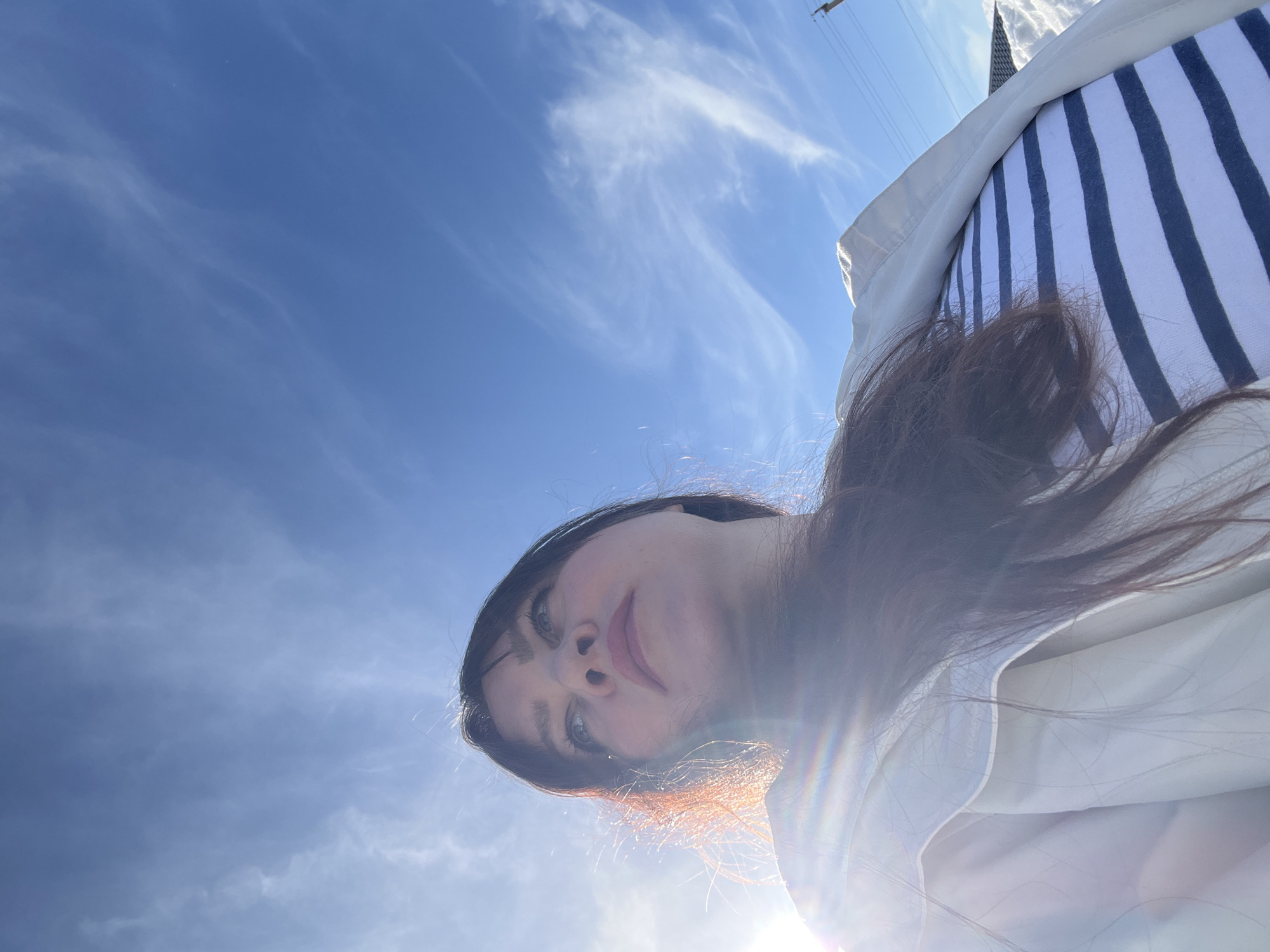 A low-angle upward photograph depicts a female individual outdoors in Noviy, Russia, on a bright, sunny day. The subject, positioned centrally, has dark hair with sun-highlighted reddish tones and is looking upwards and to the right. She is wearing a light-colored, possibly white or cream, jacket or coat over a white top with dark horizontal stripes. Strong sunlight emanates from the lower left, creating lens flare across the image. The upper two-thirds of the frame consist of a clear blue sky with sparse, wispy white clouds. In the far right background, a utility pole with wires is visible, and below it, a mound of snow suggests a cold-weather environment.