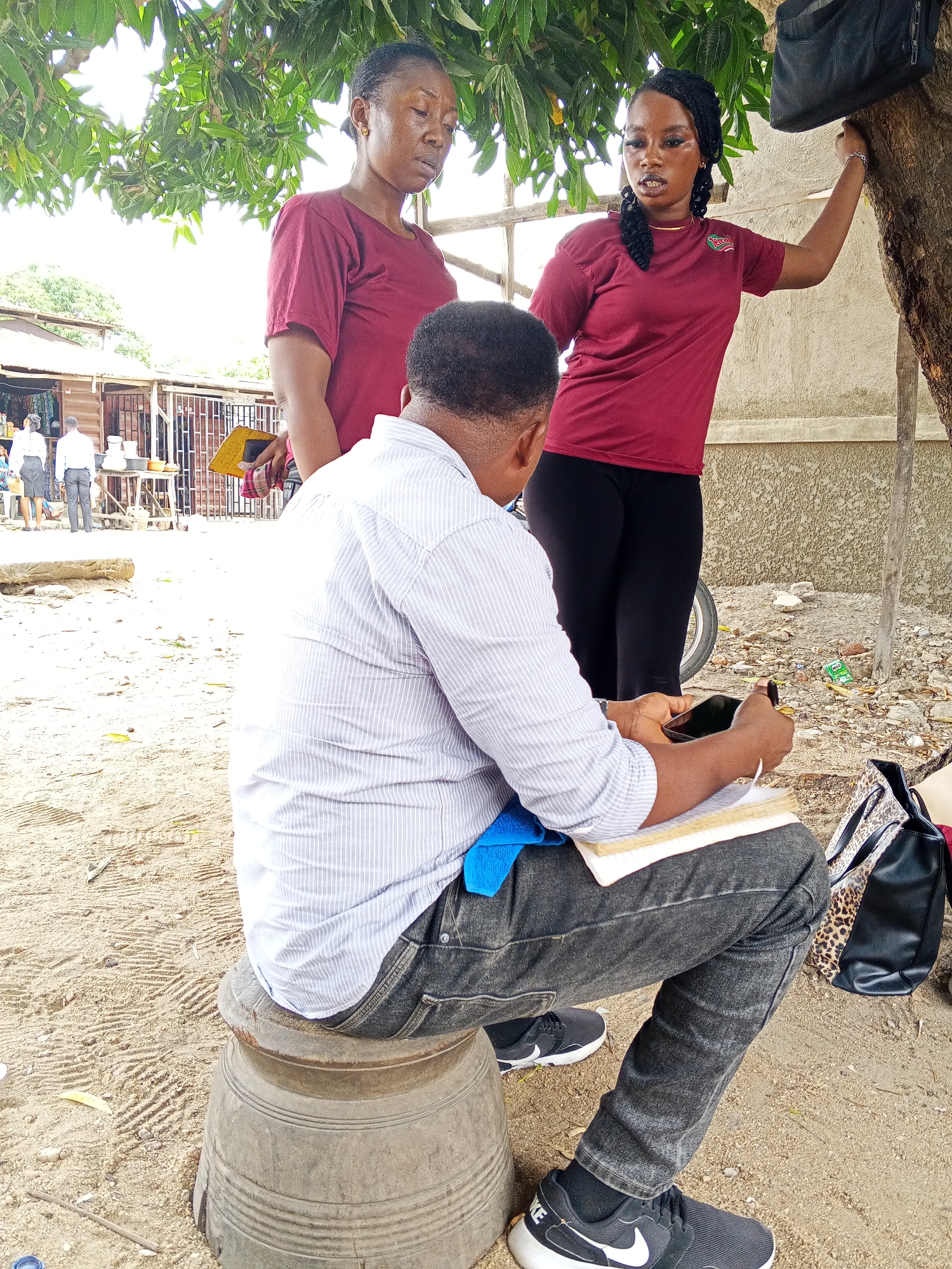 The image shows two women and a man in what appears to be an outdoor location in Zango-ndaji, Nigeria. The man is sitting on a large, gray, cylindrical object with horizontal ridges, possibly a mortar, holding a book and a cell phone in his hands. He wears a striped shirt, dark jeans, and black Nike sneakers with white accents. One woman stands behind him to the left, wearing a maroon shirt and holding a yellow object in her right hand. The other woman stands to the man's right, wearing a maroon shirt, black leggings, and black hair. Her right hand is touching the tree to her right. A tree with green leaves provides shade above the group. In the background, there's a building, tables, and several other people wearing white shirts. The ground is sandy with scattered debris. A black leather purse and an animal-print bag are on the ground to the right of the man.