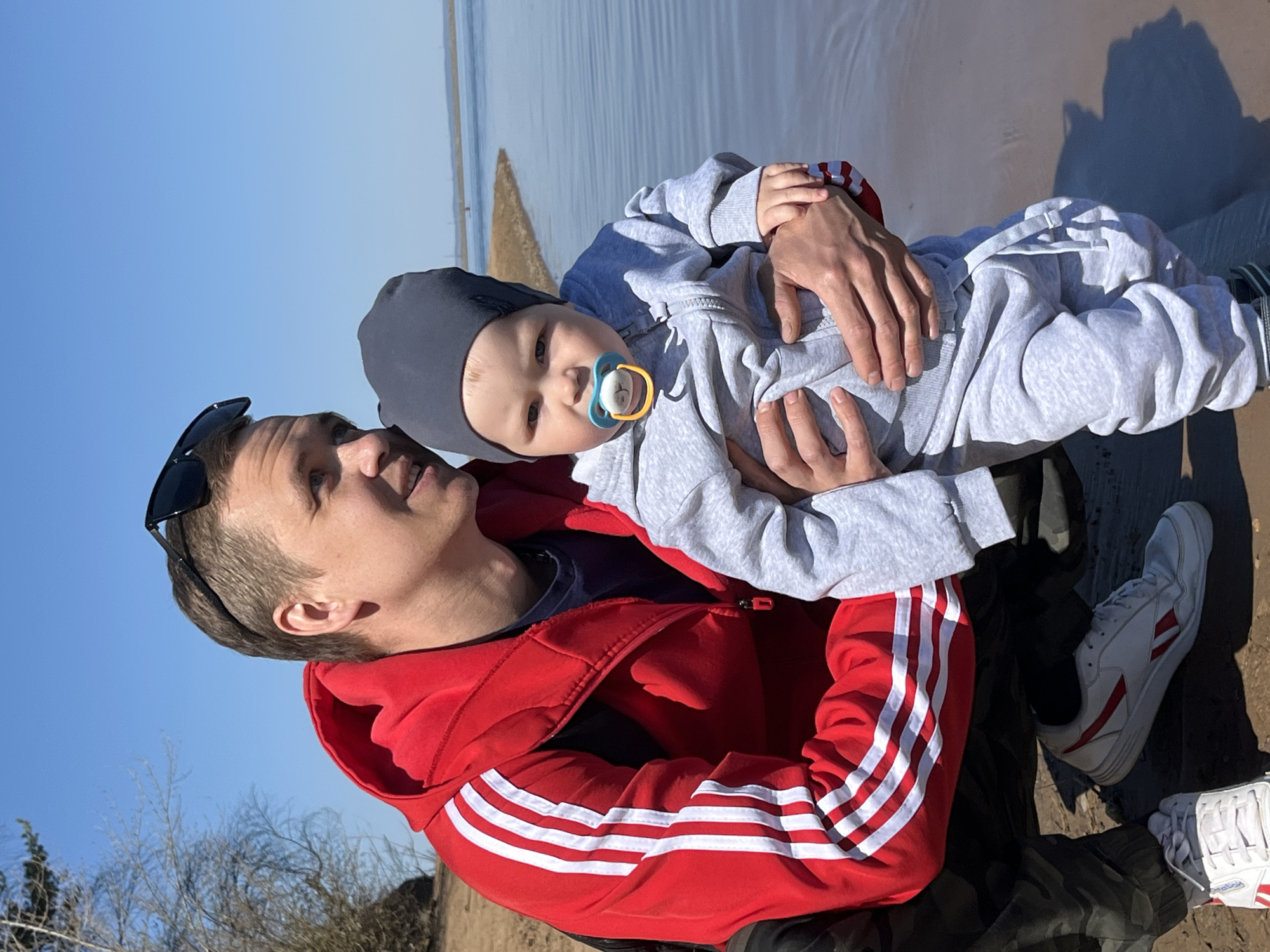 A man is holding a baby on a beach in Chaikovsky, Russia. The man is wearing a red Adidas jacket with white stripes and sunglasses on his head. The baby is wearing a grey hooded jumpsuit and a grey hat, with a pacifier in its mouth. The background shows a beach with water and sand. The sky is blue. The man is looking at the baby.