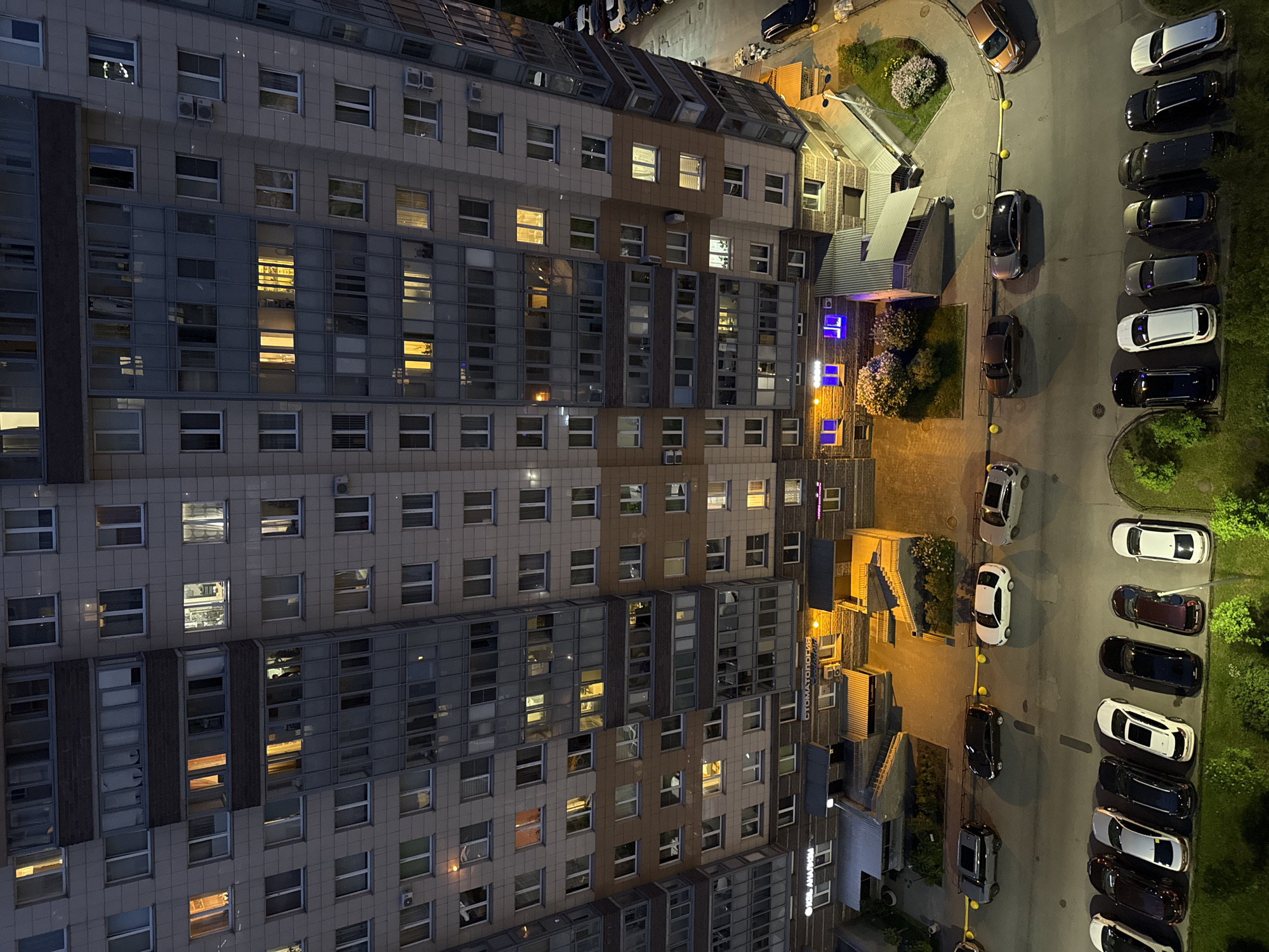 An aerial, high-angle view depicts a multi-story residential building and its immediate urban environment at night in Unknown, Russia. The building exhibits a complex modern facade with varying architectural depths, primarily light-colored panels, and interspersed sections of brown brickwork and glassed-in balconies. Numerous windows throughout the structure are illuminated from within, indicating occupancy. Ground-level commercial spaces are visible, including a "СТОМАТОЛОГИЯ" (Dentistry) clinic marked by a brightly illuminated yellow awning and entrance stairs, and another establishment labeled "ОКБЛ АНАЛИЗЫ" (Medical Tests). Distinct blue-purple artificial lights emanate from a ground-level alcove. The immediate foreground features an asphalt parking area where multiple vehicles are parked in rows; these include various sedans and SUVs of differing colors, such as black, white, silver, and dark grey. Yellow bollards delineate segments of the parking and pedestrian zones. Small landscaped sections with green grass and shrubs, some bearing light-colored blossoms, are situated around the building's base and along the parking lot perimeter. Artificial lighting from within the building and exterior fixtures provides illumination for the scene.