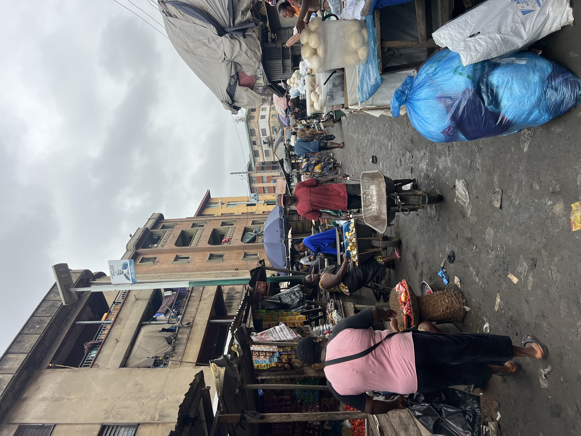 The image depicts a busy street market in Nkpor, Nigeria, under a cloudy sky. In the foreground, a person wearing a pink t-shirt and dark pants walks away from the camera, carrying a black trash bag in their left hand and a woven basket with food items on their right hip. To their right, a woman in a patterned dress sits on a stool next to a cart filled with round, pale objects, possibly food items like yam or bread. A man in a red t-shirt and a cap walks towards the camera, pushing an empty wheelbarrow.
The market is lined with makeshift stalls constructed from wood and corrugated metal. Various goods are displayed, including packaged snacks, bags of produce, and what appear to be sacks of rice or other staples. Several people are visible in the background, engaged in shopping or selling. Buildings with multiple stories, some appearing weathered, rise behind the market stalls, showcasing balconies and laundry hanging out to dry. A street light pole with a campaign banner is also visible. The ground is dirt and appears damp and littered with debris. Large blue and white plastic bags, likely containing goods or trash, are prominently placed on the right side of the frame.