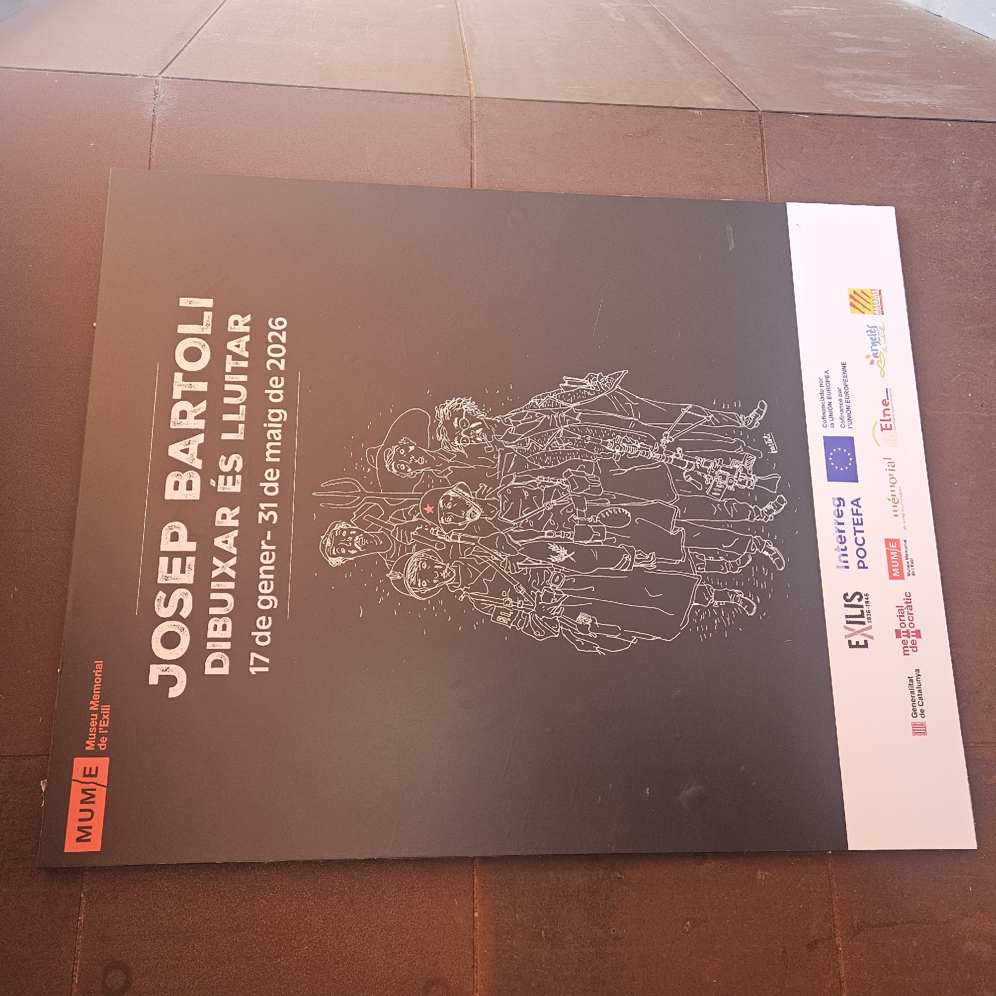 The image shows a poster advertising an exhibition of Josep Bartoli's work, titled "Drawing is Fighting," running from January 17 to May 31, 2026. The poster has a black background with the exhibition details in white text, and features a line drawing of a group of armed individuals. The bottom right of the poster includes logos of supporting organizations, including the Generalitat de Catalunya, Memorial Democràtic, Interreg POCTEFA, and others. The poster is placed on an outdoor surface, likely a paving or stone area. The location is indicated by the logo of MUME, the Museu Memorial de l'Exili (Exile Memorial Museum), located in La Jonquera, Spain.