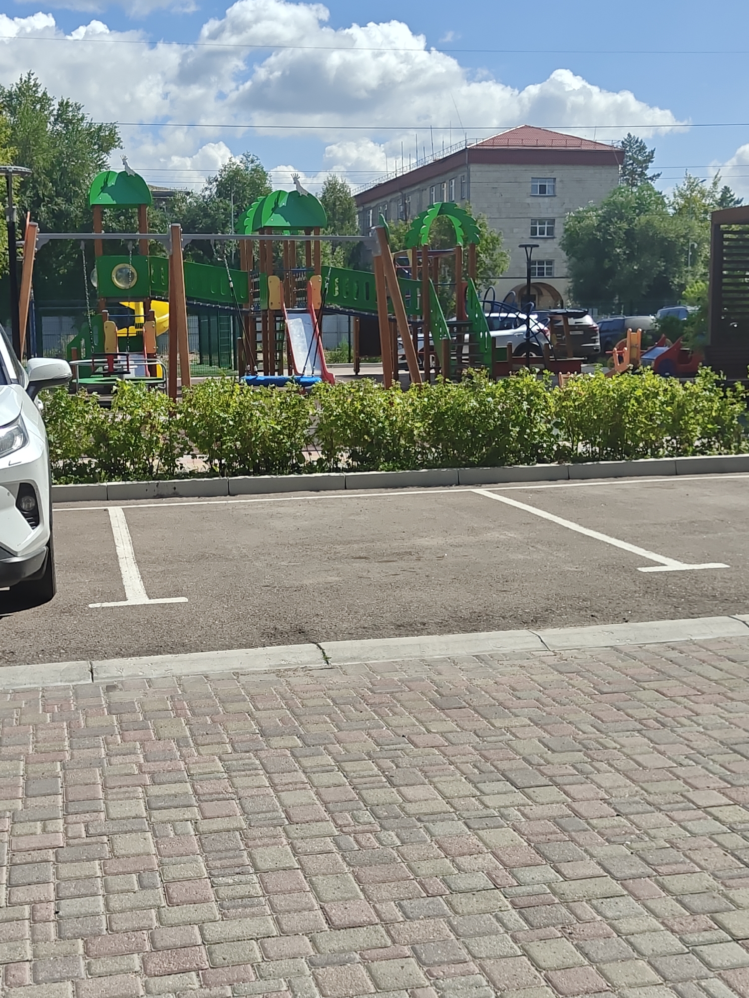 The image depicts a paved parking lot in front of a playground. A white vehicle is partially visible on the left side of the frame. The playground features green and wooden structures, including slides and swings. Beyond the playground, a multi-story building with a red roof is visible, along with trees and a cloudy blue sky. The foreground is a section of brick paving stones. The scene appears to be located in Petropavl, Kazakhstan, as indicated by the context provided. There are no people visible in the image.