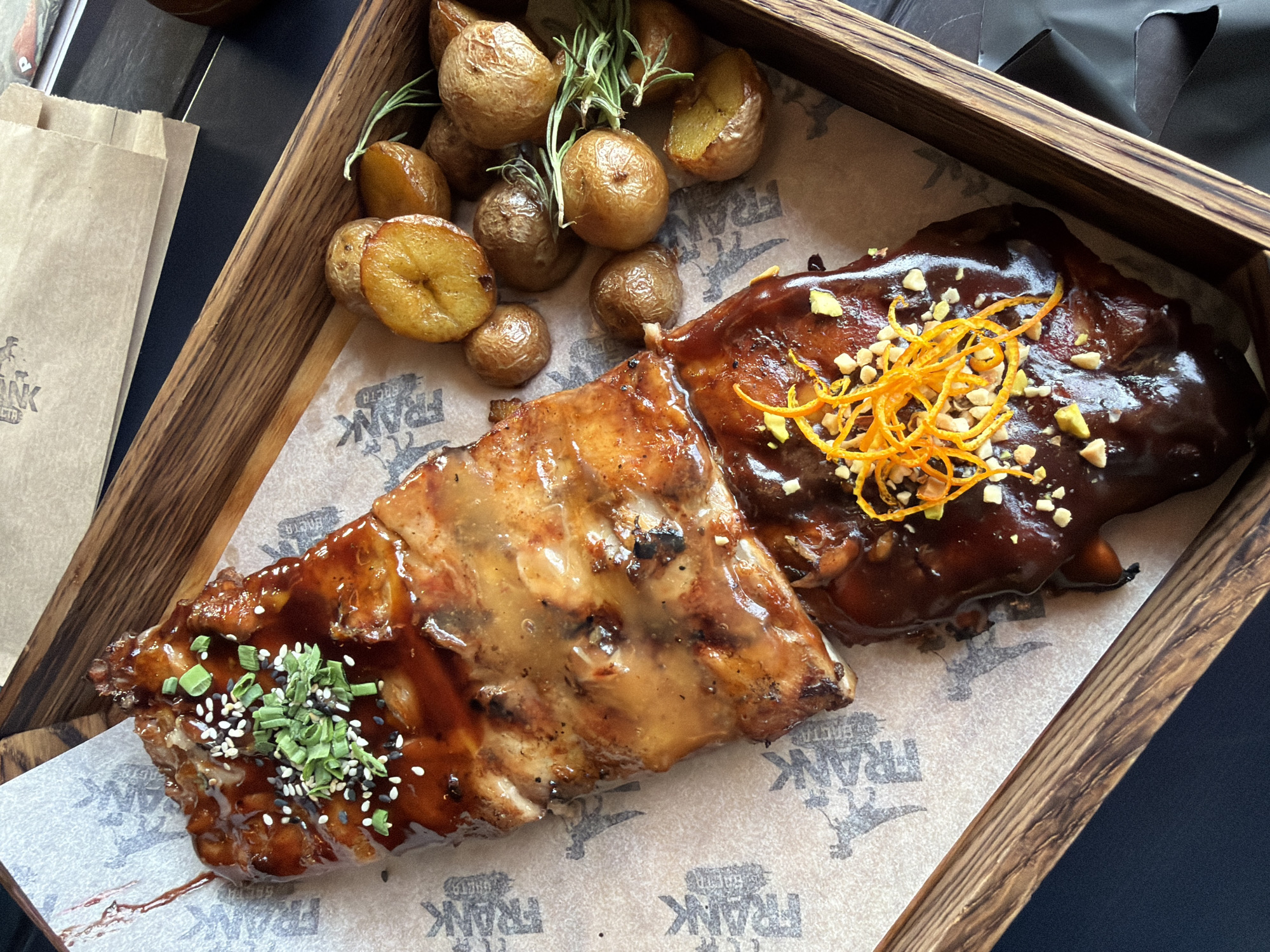 This close-up, high-angle shot presents a hearty meal served in a rectangular wooden tray. The main subjects are two large sections of barbecued ribs, roasted baby potatoes, and fresh rosemary sprigs, all resting on parchment paper.
The ribs are prepared with two distinct glazes. The left portion features a glossy, lighter reddish-brown sauce, garnished with finely chopped green onions and a sprinkle of both white and black sesame seeds. The right portion is coated in a darker, richer, and thicker sauce, topped with bright orange zest or thin strips and scattered pieces of light-colored, chopped nuts, possibly pistachios or peanuts.
Accompanying the ribs, a generous serving of whole and halved roasted baby potatoes, golden-brown and slightly glistening, fills the upper part of the tray. Several sprigs of fresh green rosemary are nestled among the potatoes, adding an aromatic and visual touch.
The parchment paper lining the tray features a repeating design, prominently displaying the word "FRANK" in a distinctive font, alongside a stylized emblem that appears to be a figure or animal. The Cyrillic text "ФРАНК" is also visible on the paper.
In the upper left background, a brown paper bag is partially visible, suggesting a take-out or restaurant setting, adjacent to a dark, untextured surface. The lighting is bright and even, highlighting the textures and glossy finishes of the food. The scene depicts a well-prepared meal, likely offered at an establishment in Krasnoyarsk, Russia.