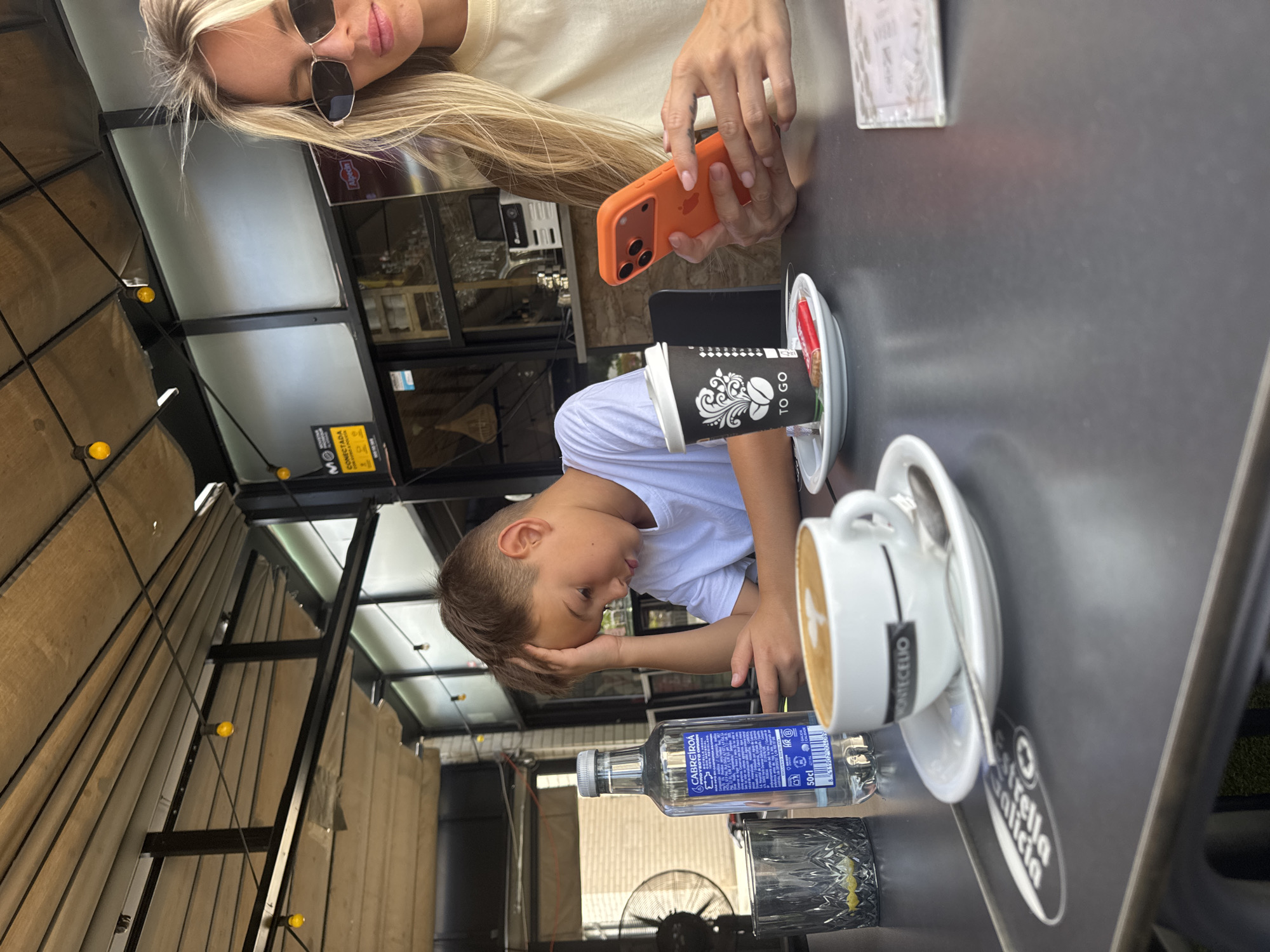 In the city of la Vila Joiosa, Spain, a boy sits at a table with his head in his hand, appearing distressed, while a woman to his right holds an orange iPhone and appears engaged with her phone. The table is set with a bottle of Cabreiroá water, a glass with ice and lemon, a coffee cup with latte art, a "To Go" coffee cup, and a small cookie. In the background, a fan is visible, along with a cafe setting featuring a bar and outdoor seating under a covered patio.