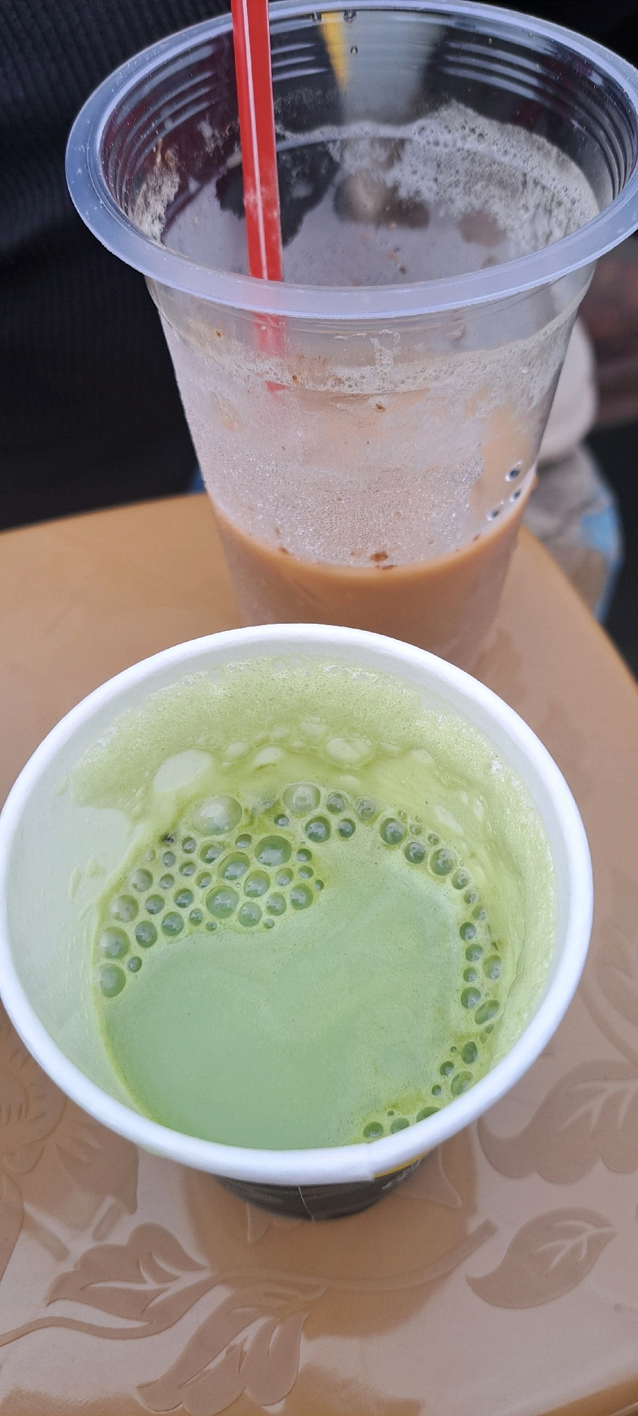 In Sumber Gedang, Indonesia, two drinks sit on a patterned table. In the foreground, a white paper cup contains a vibrant green, frothy beverage, possibly matcha. The drink is topped with numerous small bubbles. Behind it, a clear plastic cup holds a light brown, frothy liquid, with a red straw inserted. The plastic cup has condensation on its exterior. The table surface has an embossed floral design. The background is blurred, showing dark fabric and hints of other colors, suggesting an outdoor or casual setting.