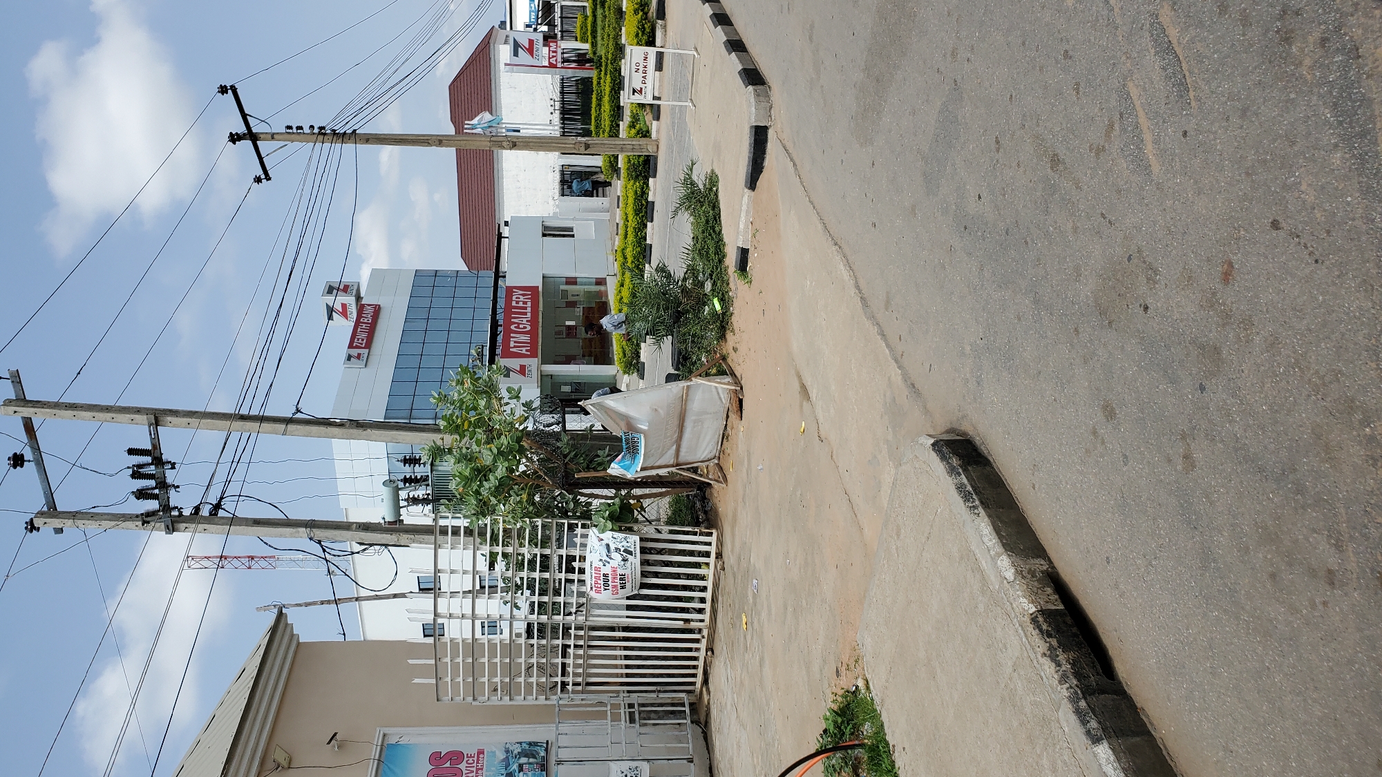 The image depicts the exterior of a Zenith Bank branch, specifically an ATM gallery, in Jalingo, Nigeria. The foreground shows a paved street with a concrete curb and a drain. A white sign with black lettering is visible on the right side of the curb, indicating "NO PARKING". To the left of the street, there is a sidewalk with a white metal gate. A white, temporary barrier with blue and white signage is placed near the gate. Beyond the gate, a building with a white facade and glass windows is visible, with the Zenith Bank logo and "ATM GALLERY" signage. Another building with a red roof and white walls is situated behind the Zenith Bank branch. Several electrical poles with wires crisscross the sky. A few people are present near the ATM gallery entrance and within the bank premises. The sky is partly cloudy. The overall scene suggests a commercial area in Jalingo.
