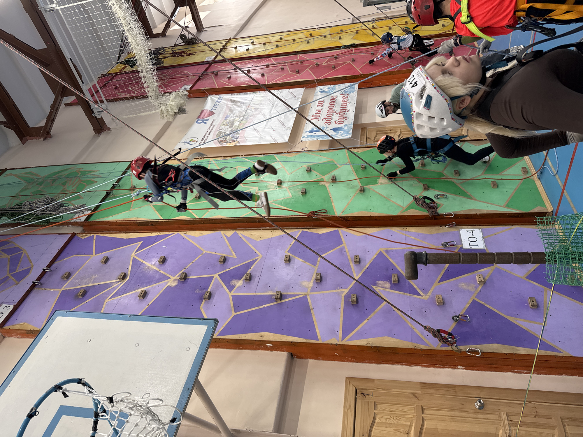 The image shows an indoor climbing wall with several people climbing. The wall is divided into sections of different colors, including purple, green, red, and yellow. Each section has a pattern of handholds and footholds. The climbers are wearing helmets and harnesses and are connected to ropes for safety.
In the foreground, there's a basketball hoop. Banners are hung above the wall, and ropes are seen crossing the frame. The location appears to be indoors, possibly in a gymnasium or recreation center. The city is Unknown, Unknown.
