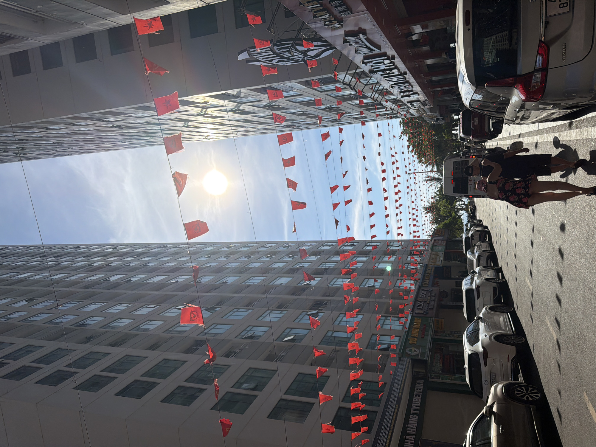 In the city of Bac Nha Trang Ward, Vietnam, a street scene unfolds under a bright, sunny sky. Two tall, modern buildings flank a street lined with parked cars. Red flags, strung across wires, flutter in the breeze, decorating the scene. The buildings appear to be residential or hotel complexes, with numerous windows visible on their facades. The street is paved, and white lane markings are visible. Several cars are parked along the side of the street, including a white SUV and a gold sedan. A couple is walking away from the camera, down the street. The man has his arm around the woman. The scene suggests a commercial or tourist area, with shops and businesses visible on the ground floor of the buildings. The sun is high in the sky, casting shadows on the street.