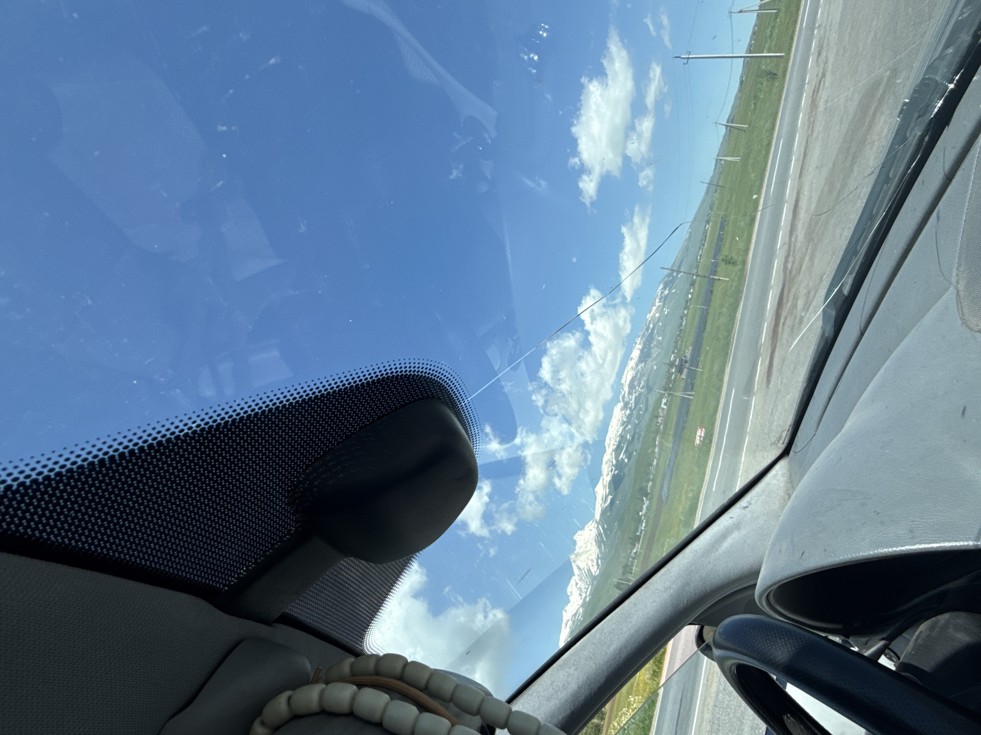 The image, taken from inside a vehicle looking through the windshield, shows a cracked windshield with a view of a landscape. Snow-capped mountains are visible in the distance, under a bright blue sky with scattered clouds. A road runs horizontally across the middle ground, with green fields and power lines flanking it. The interior of the car, including the dashboard, steering wheel, and a rearview mirror, are partially visible. There is a string of beads hanging from the interior of the car. The location is identified as Apnagyugh, Armenia.