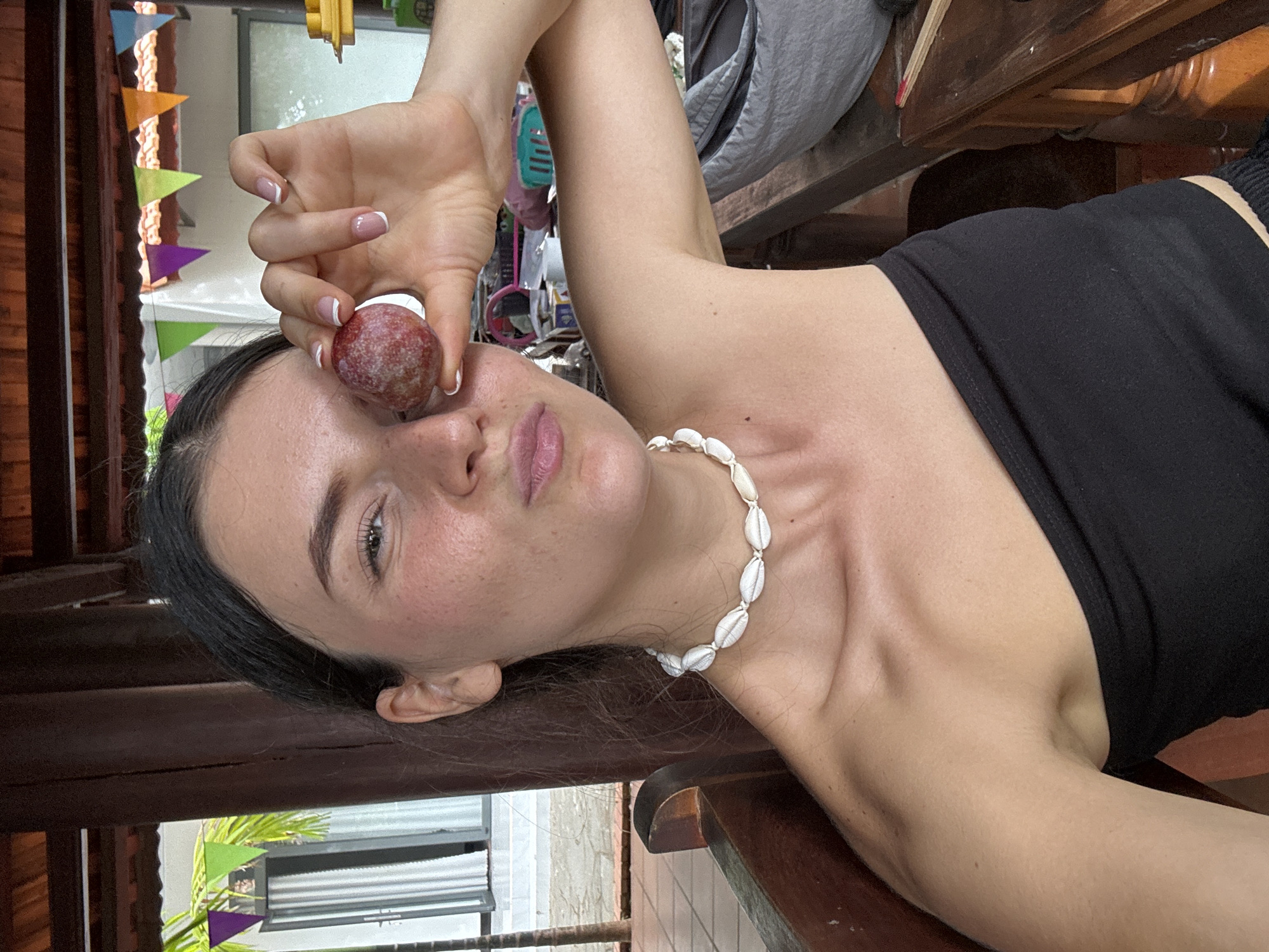 A fair-skinned woman is angled upwards, holding a small, round, reddish-brown fruit with a white base near her right eye. She is wearing a black strapless top and a seashell necklace. Her nails are manicured with a white French tip. The background shows a partially visible building exterior with a gray-slatted window covering and a leafy plant nearby. The location is likely Mũi Né, Vietnam.