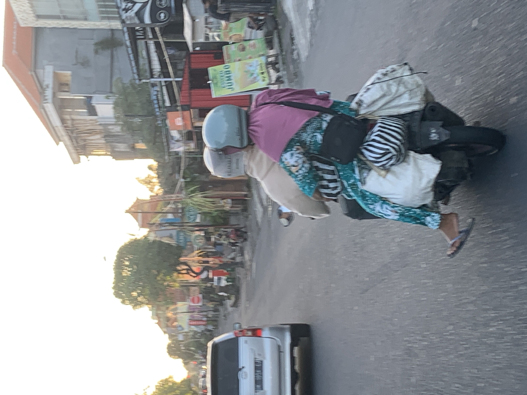 The image depicts a street scene in Denpasar, Indonesia, likely during daylight hours due to the bright sky. In the foreground, two individuals are riding a motorcycle. The person in the back is wearing a teal helmet and a pink headscarf, along with a teal and white patterned dress. They are carrying a black crossbody bag and what appears to be a zebra-patterned helmet and several bags attached to the back of the motorcycle. The person in the front is wearing a light-colored helmet and a light brown jacket. The motorcycle is on a paved road with a silver SUV visible in the left lane. The background shows buildings lining the street, including some with orange roofs, and various signs, suggesting a commercial or residential area. Trees and vegetation are also present along the roadside. The overall atmosphere is one of everyday urban life in Indonesia.