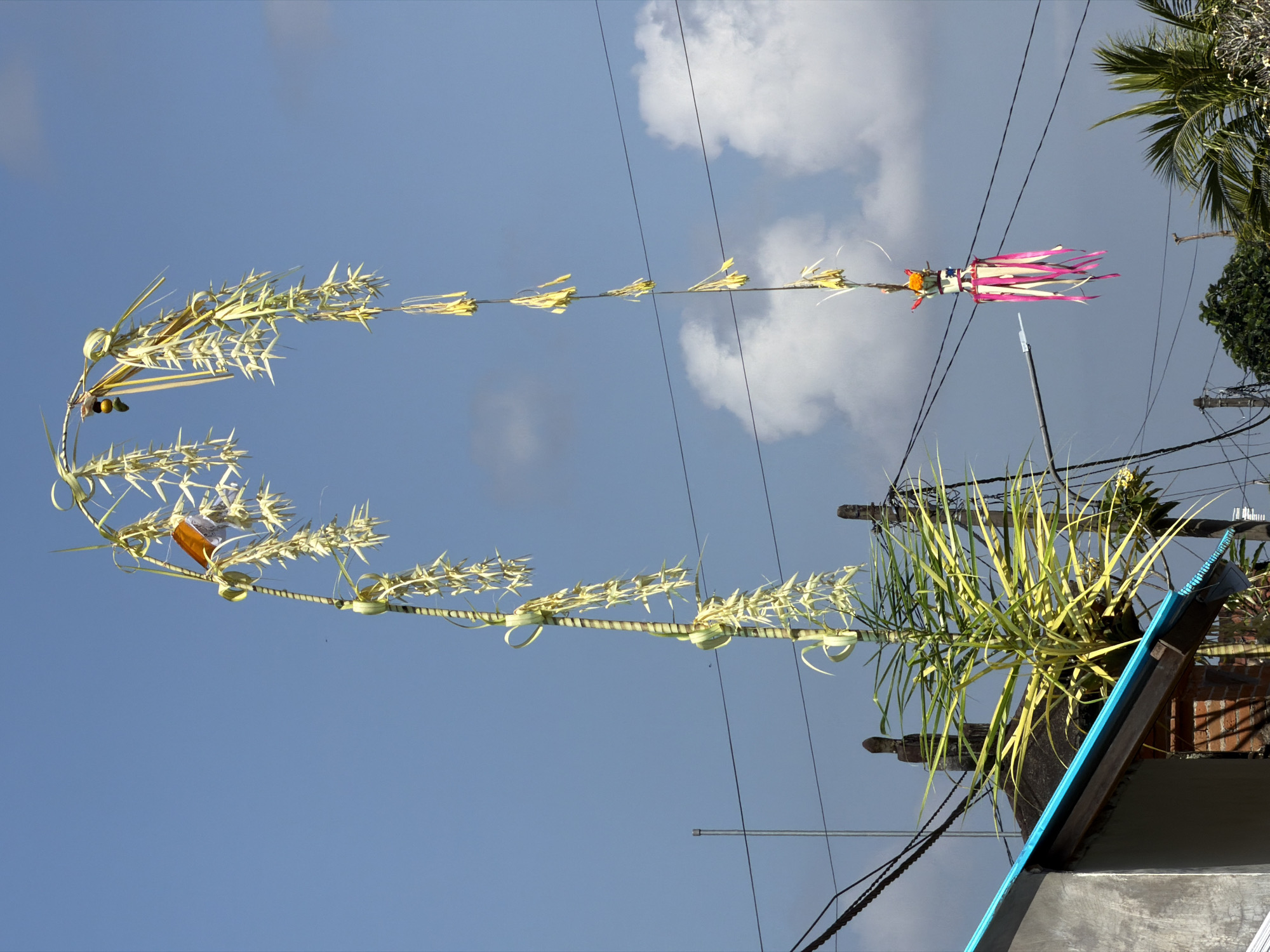 An arched bamboo pole decorated with woven palm leaves and ribbons is displayed against a clear blue sky with scattered clouds. The pole is situated in what appears to be a residential area in the Ubud District of Indonesia, as evidenced by the presence of a building with a blue-trimmed roof and electrical wires crisscrossing the background. A small, colorful offering is suspended from the lower end of the pole. The overall scene suggests a cultural or religious celebration.