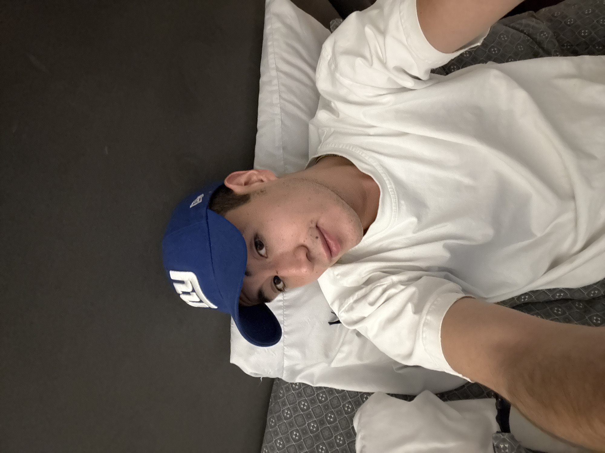 A person lies on a bed with a white pillow and patterned gray and black bedding. The person is wearing a white t-shirt and a blue baseball cap with white writing. The location in the city of San Bernardino Tlaxcalancingo, Mexico is not identifiable from the image.