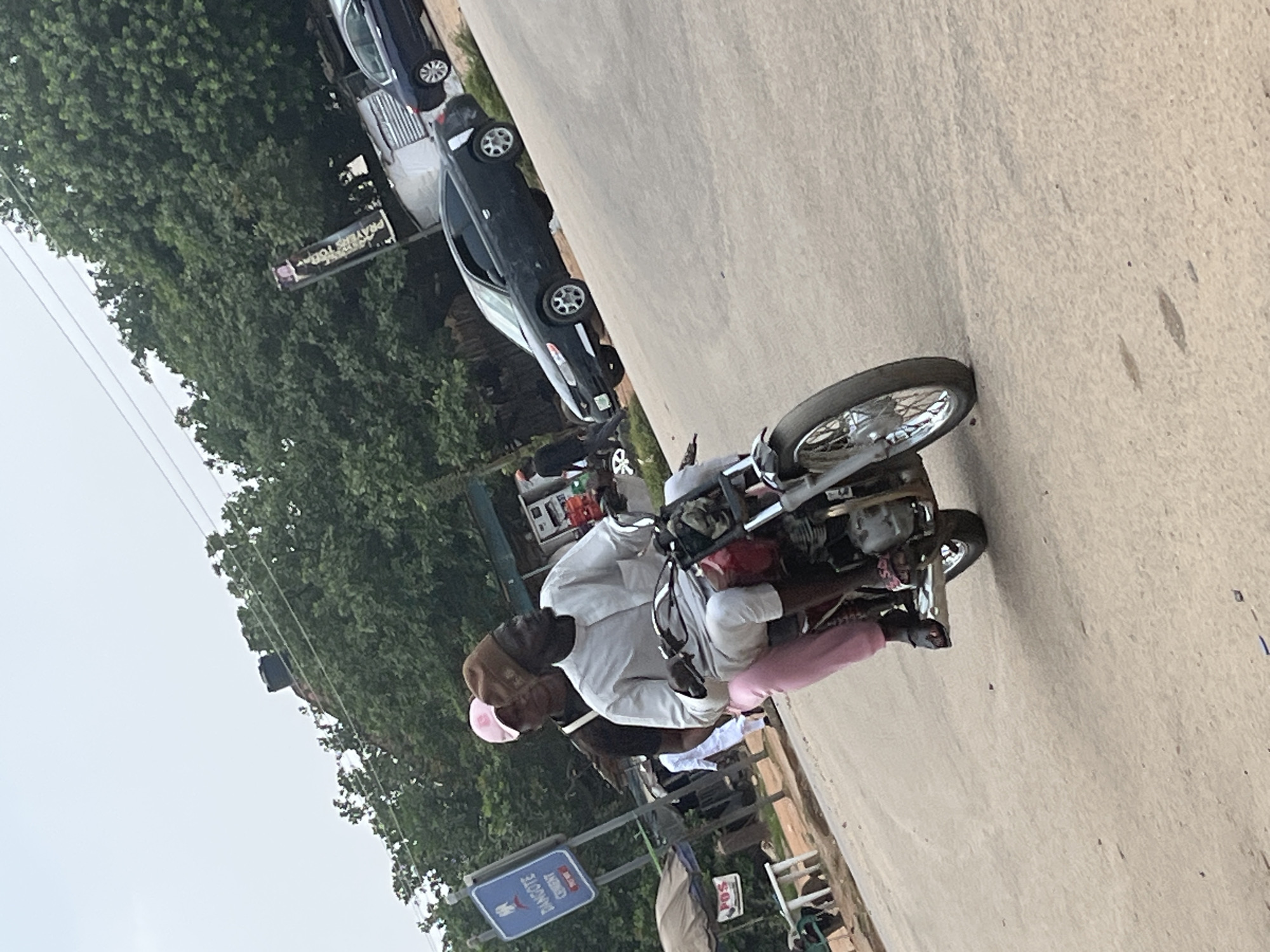 The image depicts a street scene in Abuja, Nigeria, with a focus on a motorcycle and its riders. A man, dressed in white traditional attire and wearing a brown cap, is seated on the motorcycle. He is holding onto the handlebars, with his right hand extended forward and slightly upwards. He appears to be looking downwards. Behind him, another person, wearing a pink garment, is also seated on the motorcycle, with their hands resting on the rider's shoulders.
The motorcycle is positioned on a paved road, with a light-colored concrete surface. To the left of the road, dense green foliage from trees forms a backdrop. Several parked cars are visible in the background, including a dark-colored sedan and another vehicle. A signpost with blue and white lettering is also present in the lower left corner of the image. The sign has text that appears to include "DANGOTE CEMENT".
The overall lighting suggests it could be daytime, possibly overcast given the diffused light. The scene appears to be a typical urban street environment. There are no strong indicators of specific emotions, but the posture of the riders suggests they are engaged in transit. The presence of cars and the motorcycle implies a mode of transportation in an active area.