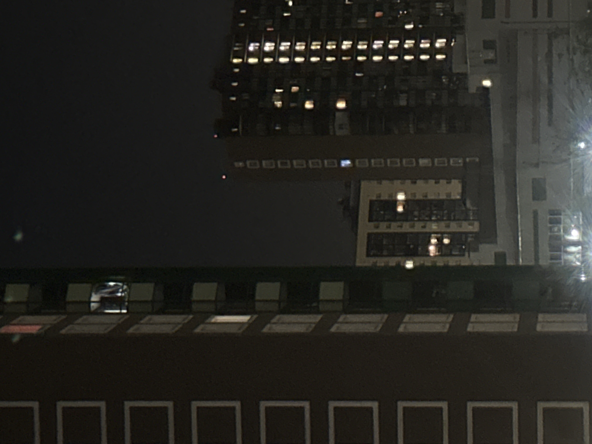 An outdoor night-time vertical medium shot captures multiple residential buildings in an urban setting. The building in the foreground on the left is dark brown with a grid of evenly spaced rectangular white frames on its facade. To the right of this building is another dark building with numerous illuminated windows, suggesting occupancy. Further in the background, to the right, is a taller, more complex building with many windows, some of which are lit, showcasing a dense urban environment at night. The sky is dark, indicating it is nighttime, and a few distant lights are visible. The overall impression is one of a densely populated urban area viewed at night.