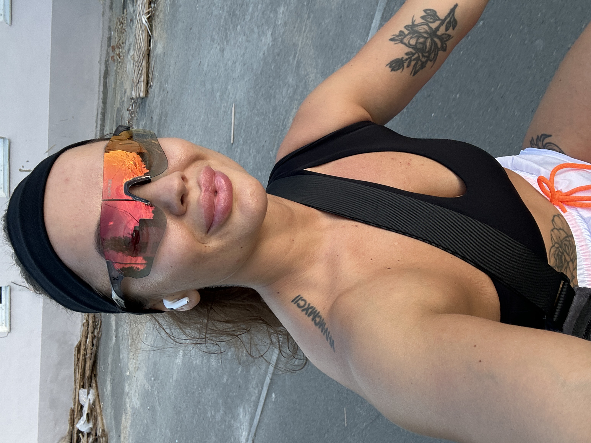 A woman is captured in a close-up, outdoor shot, her face angled towards the camera. She is wearing a black headband and mirrored sunglasses with pink lenses that reflect the surrounding environment, including buildings and a sunset. A white earbud is visible in her right ear. Her lips are slightly parted, and she appears to be outdoors on a paved surface, possibly a street. Her left shoulder is visible, showing a tattoo of Roman numerals. She is wearing a black sports bra with a strap across her chest, and white shorts with orange drawstrings. Another tattoo of a rose is visible on her right arm. The background consists of a building and piles of wooden debris. The overall impression is a casual, active moment in an urban setting.