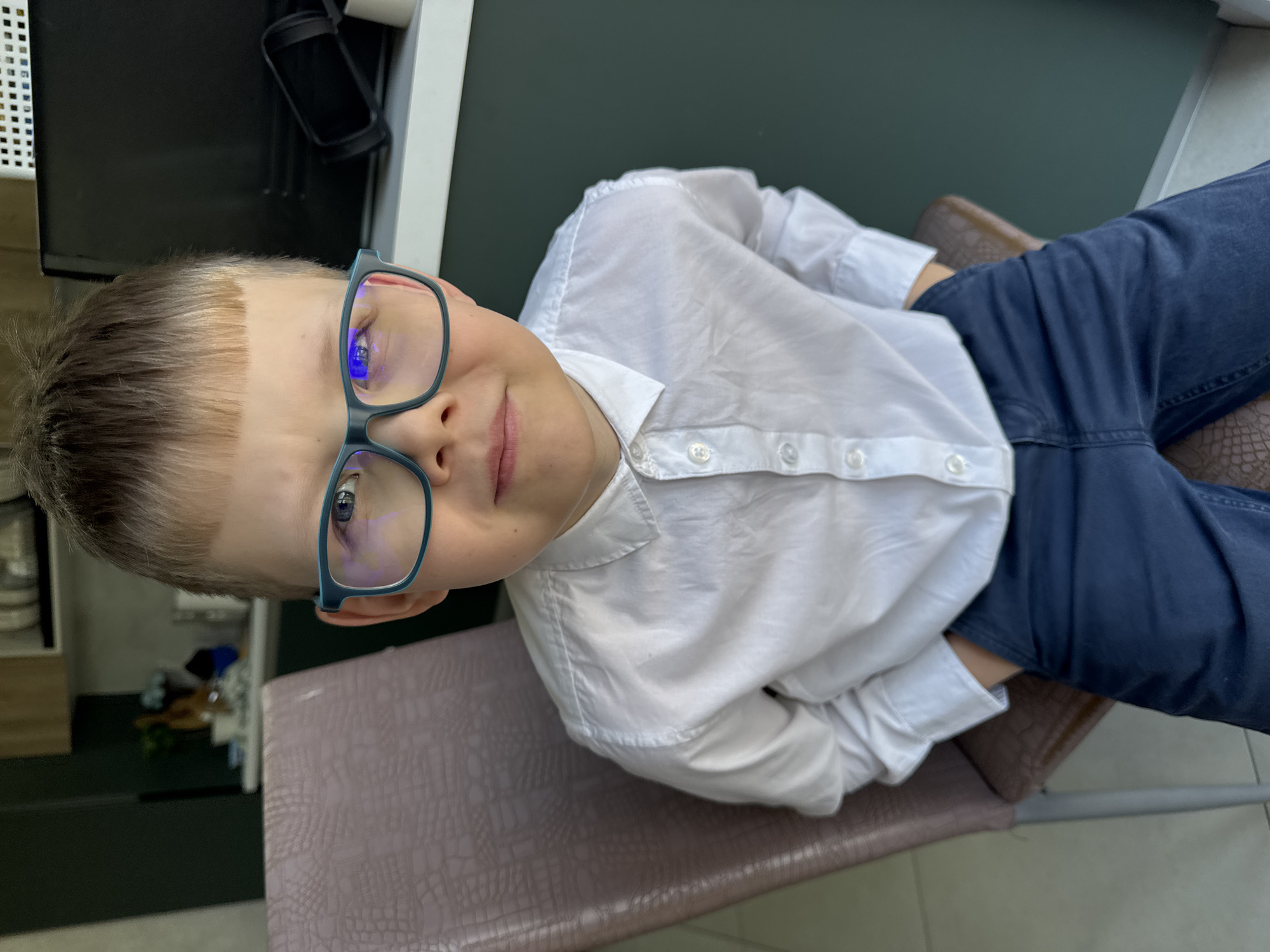 The image displays a young boy, approximately 6-9 years old, captured in a portrait shot that is rotated ninety degrees clockwise. The boy is looking directly at the camera with a gentle, slightly smiling expression. He has neatly cut, short light-brown hair and is wearing blue-framed glasses. His eyes appear light in color. He is dressed in a crisp white long-sleeved button-up shirt and dark blue trousers.
The boy is seated on a brown chair with a textured, possibly reptilian-skin-like pattern on its upholstery. The setting appears to be indoors. In the background, to the boy's left (when viewed upright), there is a dark, possibly black, piece of furniture or equipment, next to a white, gridded panel. To his right, a plain, dark green wall is visible. The lighting suggests an indoor environment, likely daytime, but no external weather conditions are discernible. No text is visible in the image. The boy's demeanor suggests he is posing for the photograph.