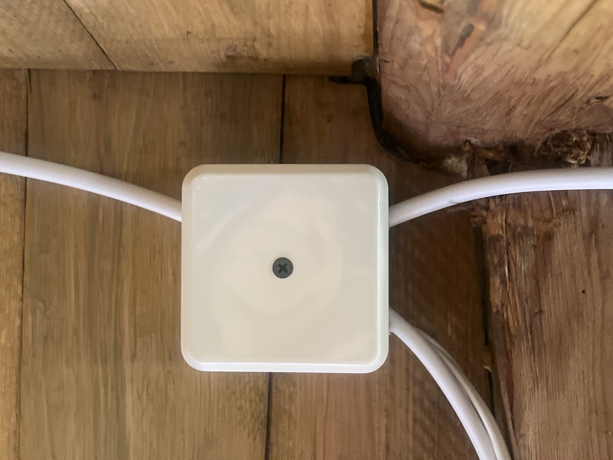 The image shows a close-up, overhead view of a white, square junction box with a single black screw in the center. Two white electrical cables enter the box from the left and right sides. The junction box is mounted on a surface of wooden planks. The background consists of rough, unfinished wooden beams and planks, suggesting an indoor or sheltered environment. The lighting is even, with no strong shadows. The scene appears to be in Norviliškės, Lithuania, likely within an older structure or unfinished building due to the raw wood. There are no people or animals visible in the image.