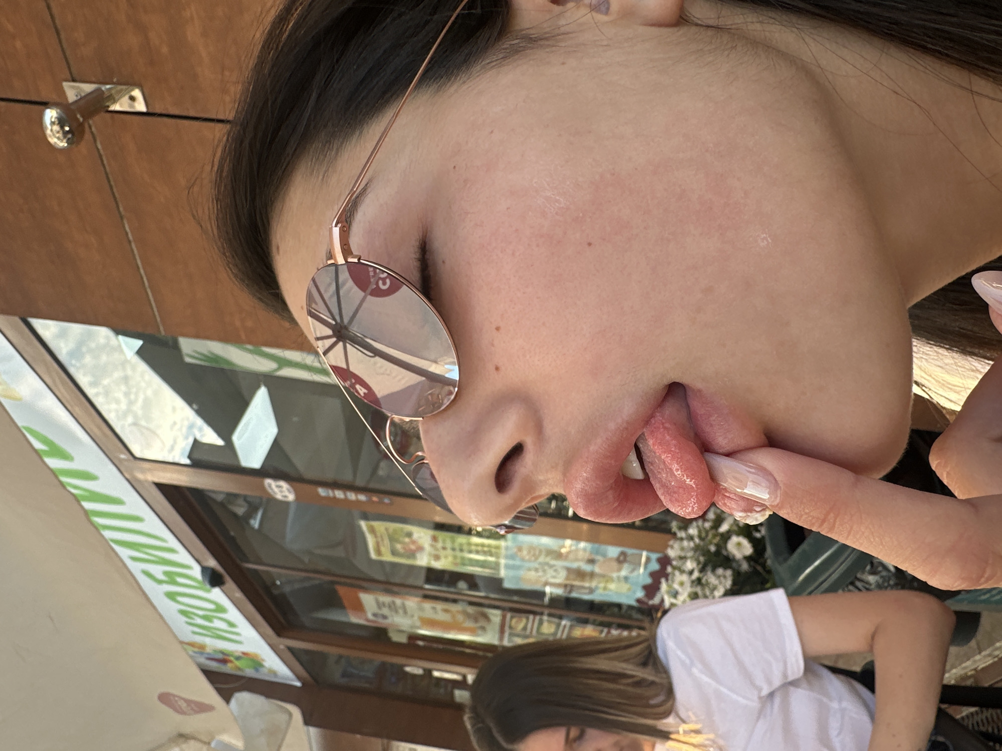 A close-up profile view of a woman wearing rose-tinted sunglasses with her tongue extended and touching her finger. Another woman is visible in the background, wearing a white shirt and with brown hair. The location is outdoors, likely a cafe or restaurant, as suggested by the door with the words "ИЗВОРИ" in Cyrillic characters on the signage. The surroundings appear to be an urban setting; based on the Cyrillic signage, it suggests the city of Plovdiv, Bulgaria.